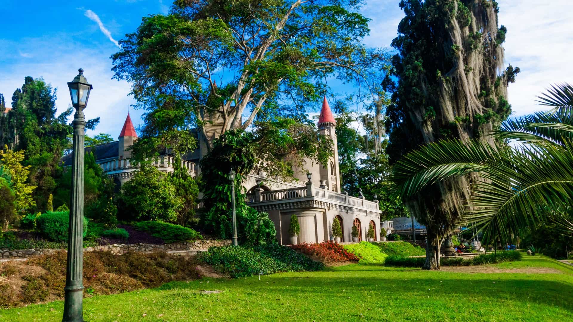 The El Castillo Museum, a mock-Gothic castle in Medellín, Colombia, stands prominently in the background, featuring stone towers and spires. In the foreground, the meticulously manicured French-style gardens are visible with lush green lawns and colorful flowerbeds, capturing a beautiful outdoor view of this elegant landmark.