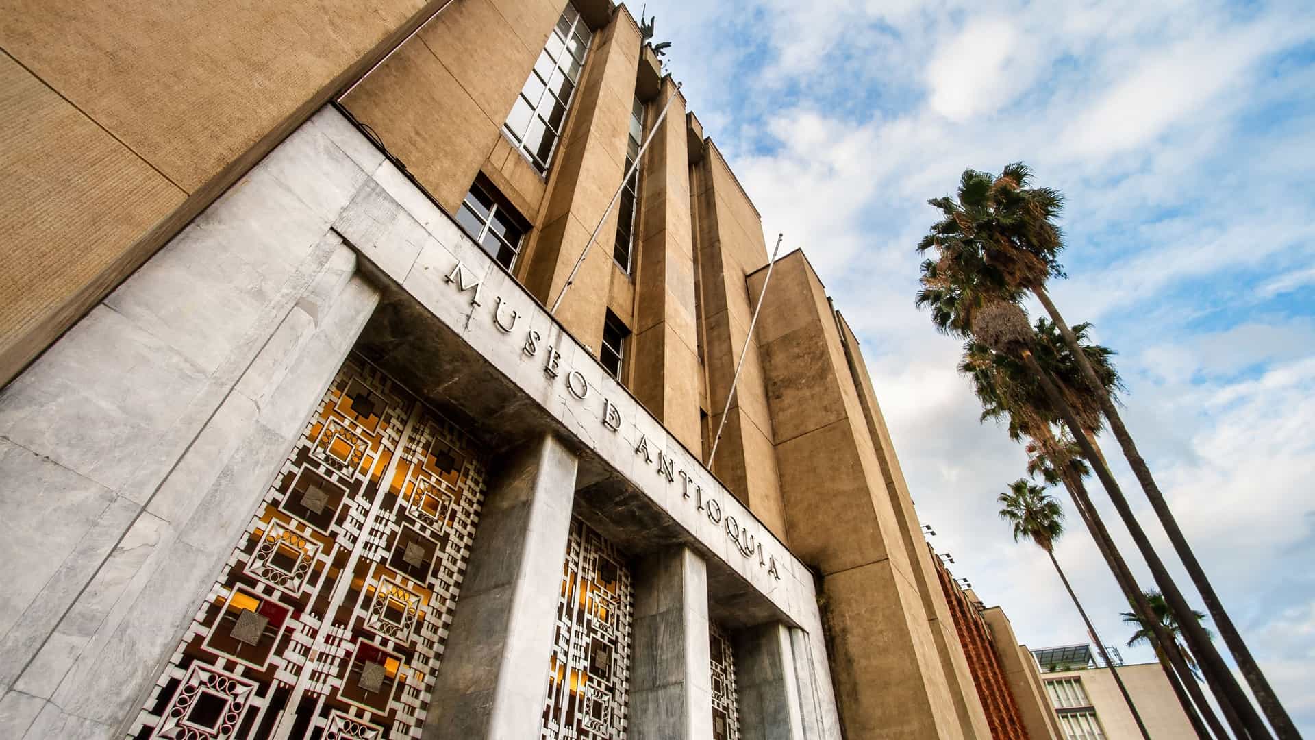 A frontal view of the facade and main entrance of the Antioquia Museum in Medellín, Colombia. The historic neoclassical building is composed of yellow-colored stone with arched windows and columns, contrasting with the dark red awning above the main entrance on a bright, sunny day.
