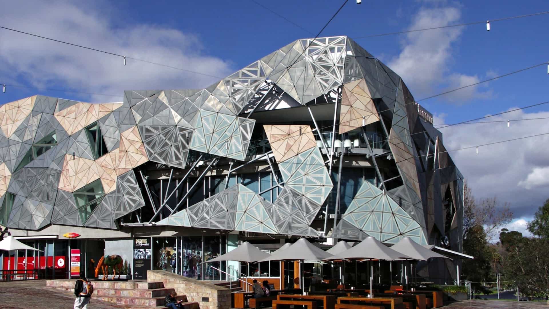 Federation Square in Melbourne, Australia, showcasing its distinctive modern architecture characterized by deconstructed, fragmented, and triangular-shaped buildings with a mix of stone, steel, and glass materials, set against a blue sky.