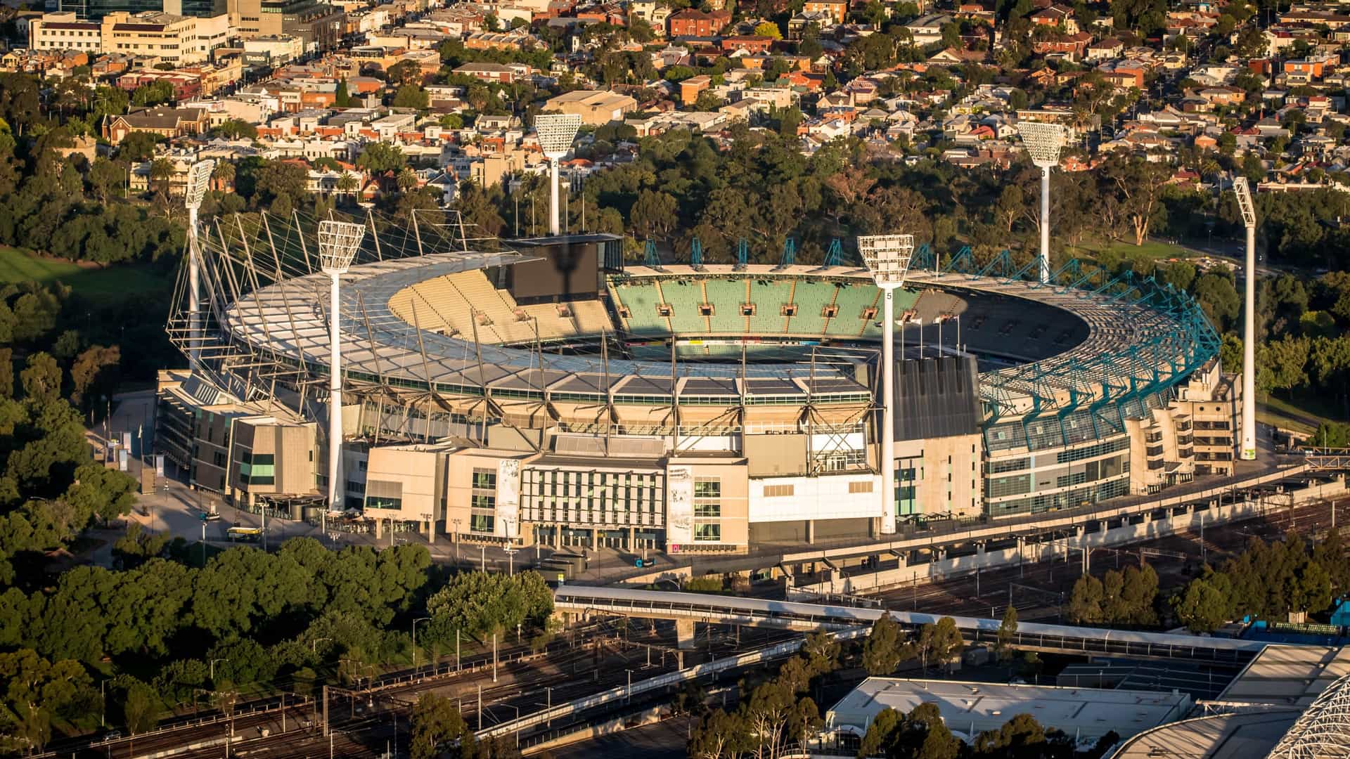 The iconic Melbourne Cricket Ground (MCG), a massive circular stadium known as "The G" and a central venue for Australian Rules Football (AFL) and Test Cricket. The image likely highlights the stadium's distinctive architecture, possibly featuring the modern stands, the colossal light towers, or the lush green oval playing field. The photo may include the vast seating bowl, or a view of the stadium complex against the backdrop of the Melbourne city skyline.