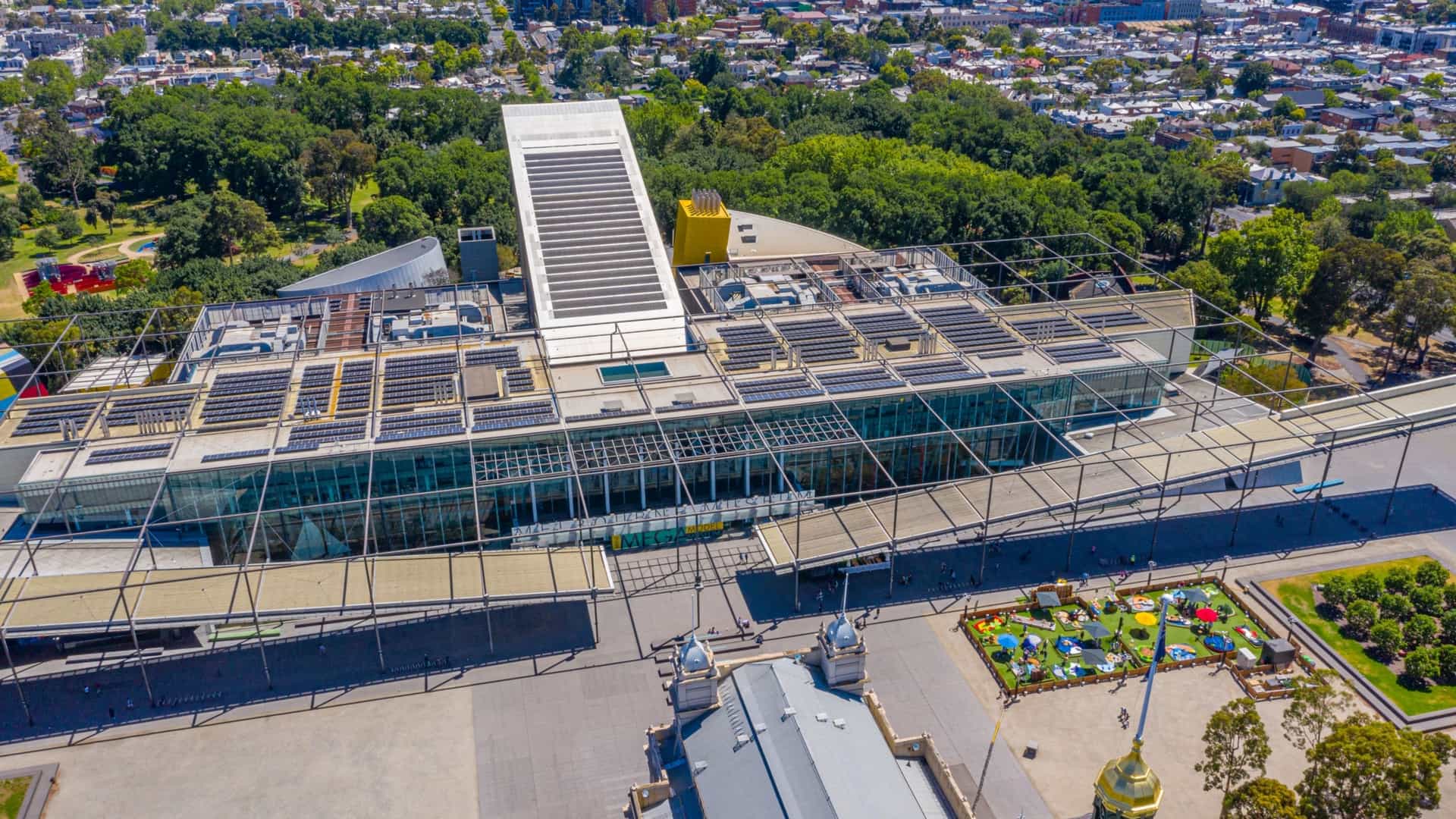 An aerial view of the modern Melbourne Museum complex and the adjacent historic Royal Exhibition Building, situated in the extensive green space of Carlton Gardens. The museum features a prominent central blade-like structure, while the surrounding city skyline of the Melbourne CBD is visible in the background.