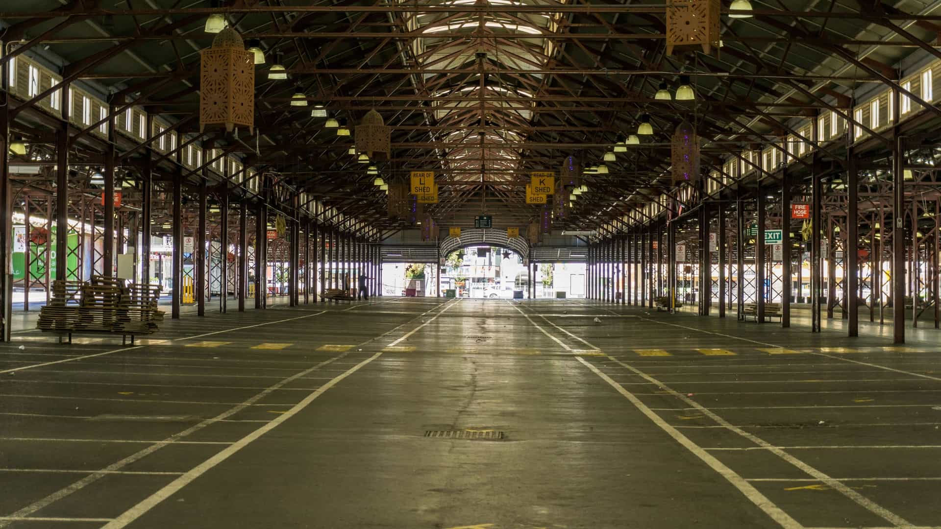 A bustling interior view of the historic Queen Victoria Market in Melbourne. The photograph shows a long aisle lined with vendor stalls, all set beneath the market's characteristic high, covered shed roof supported by visible iron trusswork and timber posts. The stalls are typically filled with merchandise such as fresh produce, deli items, or various general goods, reflecting the market's lively atmosphere.