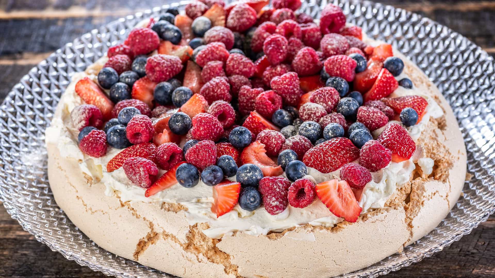 A close-up of a traditional Australian Pavlova dessert, featuring a large, baked white meringue base with a crisp exterior. The meringue is generously topped with a layer of fresh white whipped cream and garnished with a colorful assortment of fresh fruits, likely including red strawberries, green kiwi slices, and passion fruit pulp. The dessert is presented on a platter, ready to be served.