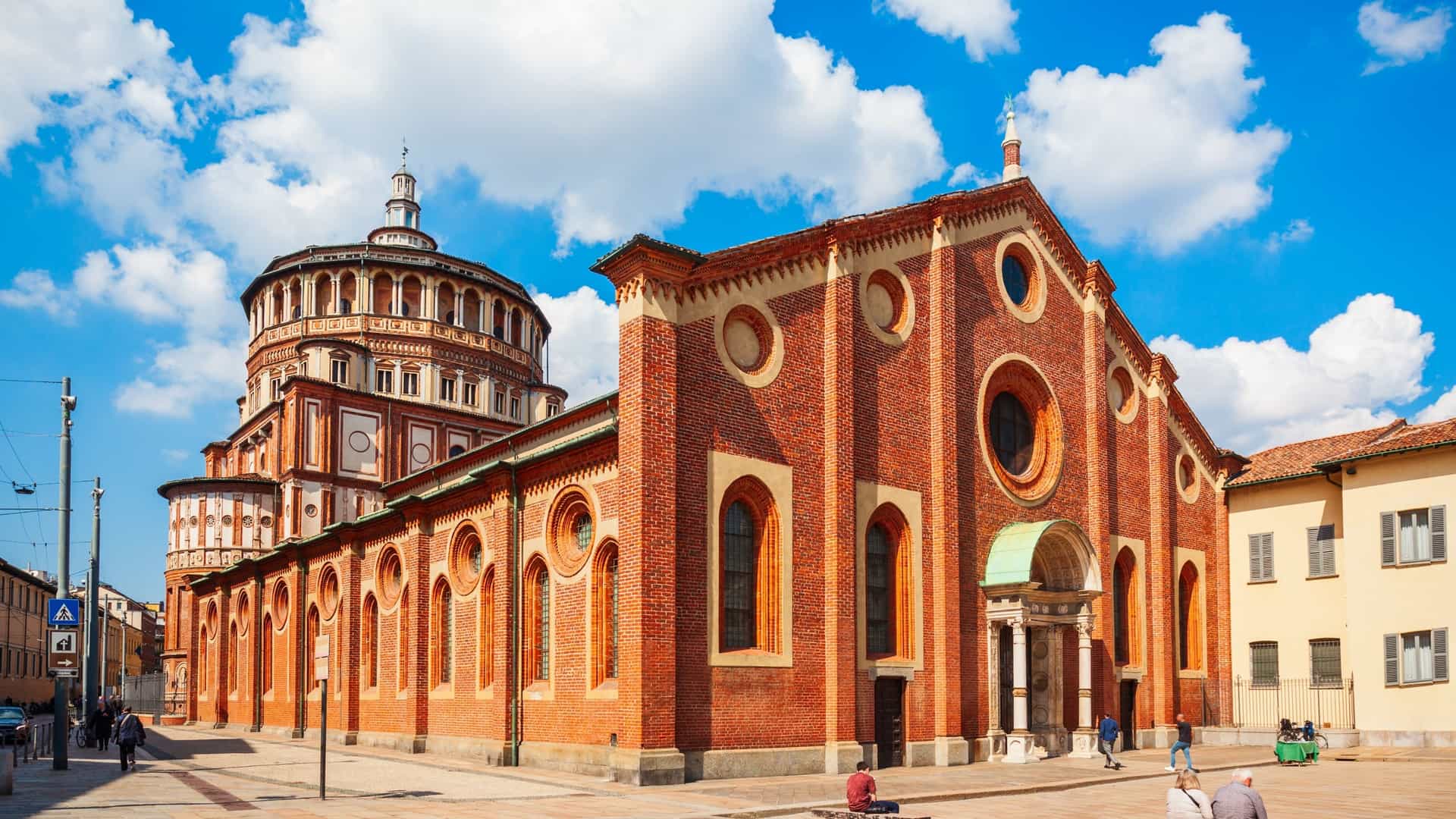A full exterior view of the UNESCO World Heritage site Santa Maria delle Grazie (Holy Mary of Graces) in Milan, Italy. The image focuses on the Renaissance-style apse, tribune, and large, sixteen-sided dome attributed to Donato Bramante, built with decorative red brick and white stone accents. This section contrasts with the simpler, gabled, Late Gothic nave of the church.