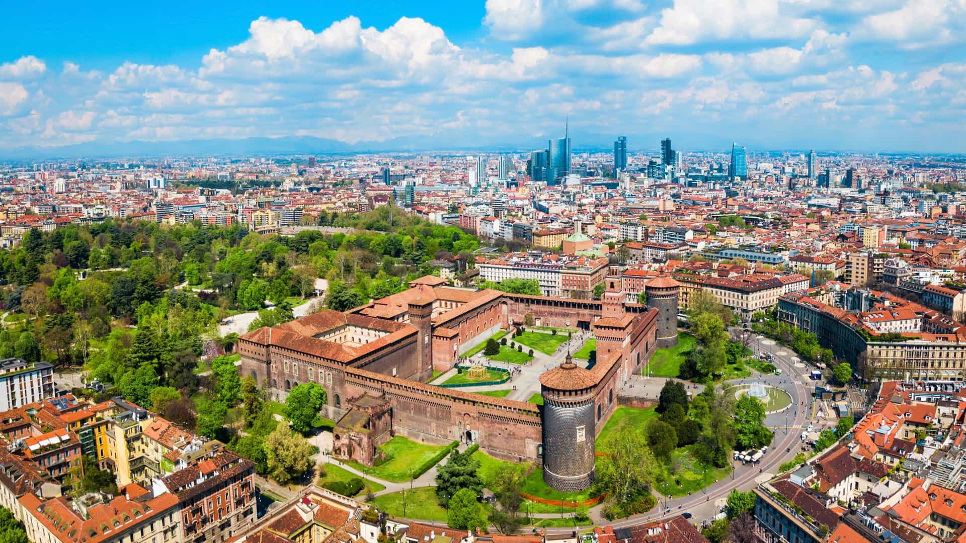  An aerial, panoramic view of the Sforza Castle (Castello Sforzesco) in Milan, Italy. The medieval fortification, built with red brick, features a large central tower, multiple smaller turrets, and extensive battlements. The surrounding Sempione Park with its trees and manicured green spaces is visible in the foreground and background. The image highlights the scale and historic architecture of the castle within the urban landscape.