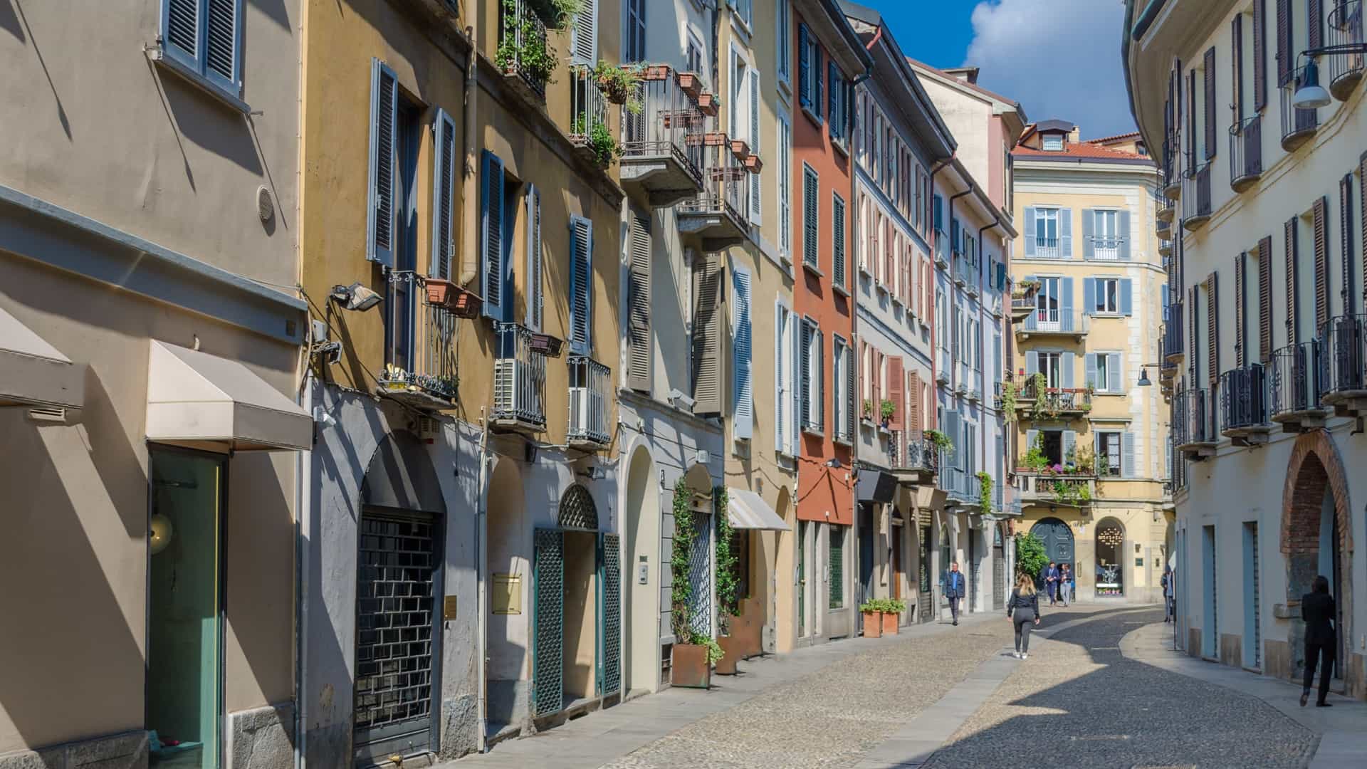 A charming, narrow, cobblestone street (likely Via Fiori Chiari) in the fashionable Brera district of Milan, Italy, lined with historic buildings. The buildings feature colorful, painted facades in shades of warm yellow and soft pink, with dark green window shutters and balconies adorned with flowering plants, conveying the bohemian and artistic atmosphere of the neighborhood.