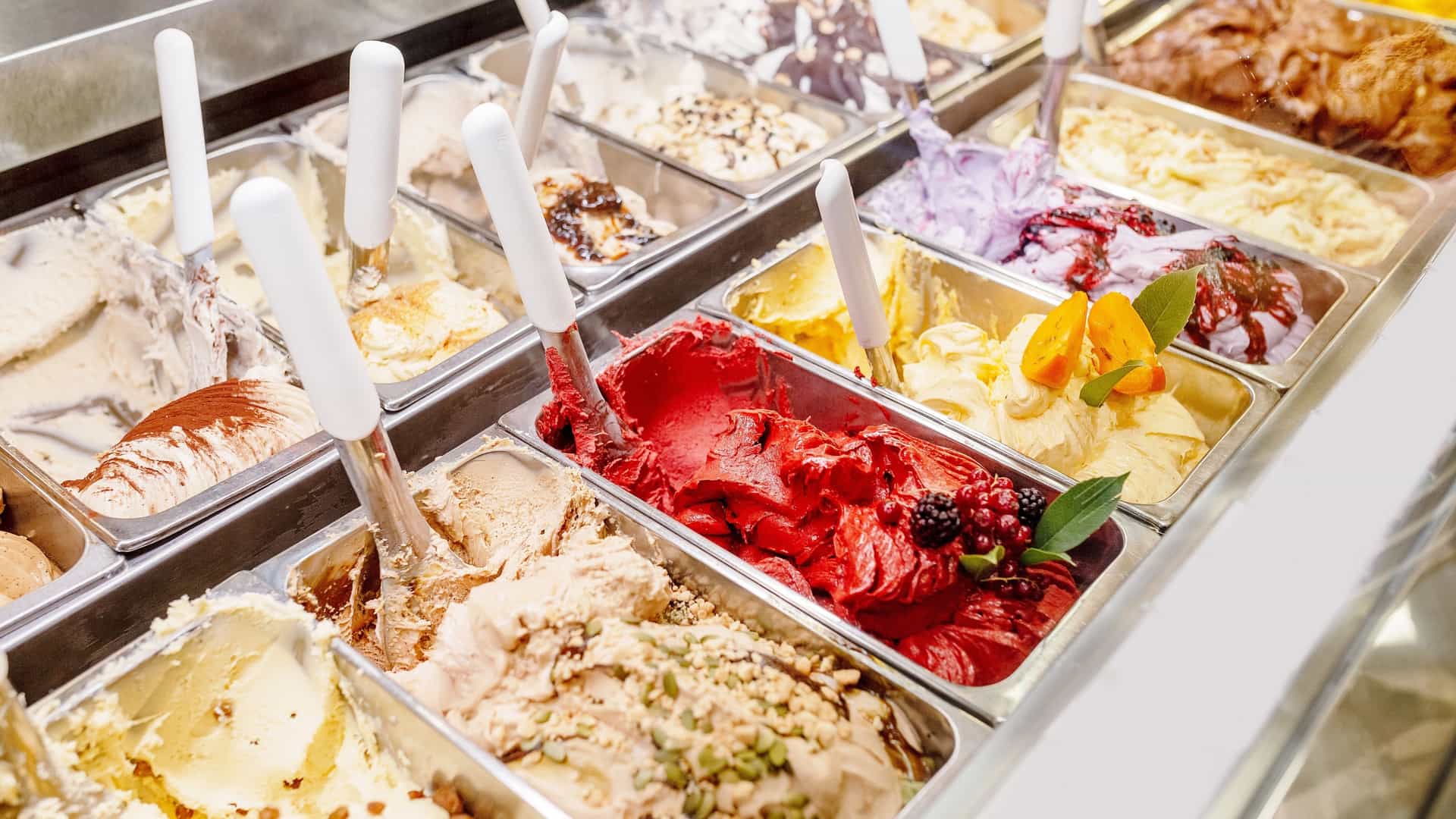  A close up, low angle view of a gelato display case in a dessert shop. The showcase is filled with various flavors of gelato, arranged in a colorful array in metal containers. The gelato has a soft, creamy texture and is mounded high in the bins. The vibrant colors range from white, brown, and yellow to bright pink, green, and red, representing a variety of fruit, chocolate, and nut based flavors.