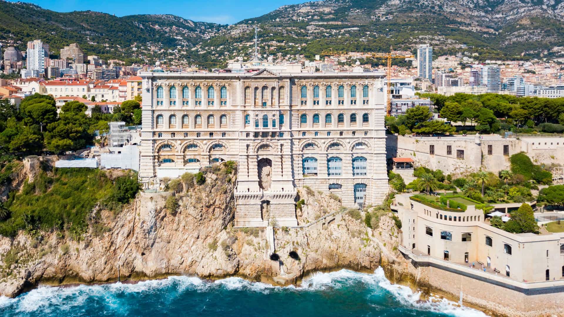The Oceanographic Museum (Musée Océanographique) in Monaco, a monumental Baroque Revival building perched dramatically on the cliffside of the Rock of Monaco, rising 85 meters above the Mediterranean Sea. The photograph is taken from a distance and shows the grand, ornate white stone facade, which includes numerous columns, arches, and sculptures, with the blue water of the sea visible below and the city of Monaco-Ville rising behind it.