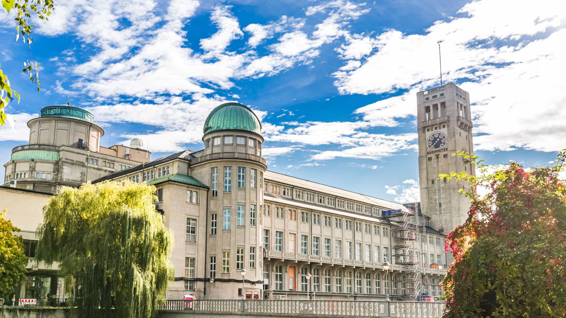 An exterior view of the historic Deutsches Museum building on Museumsinsel in Munich, Germany, situated on the Isar River, showcasing its grand architecture and the iconic tower housing a modern weather station and Germany's largest thermometer.
