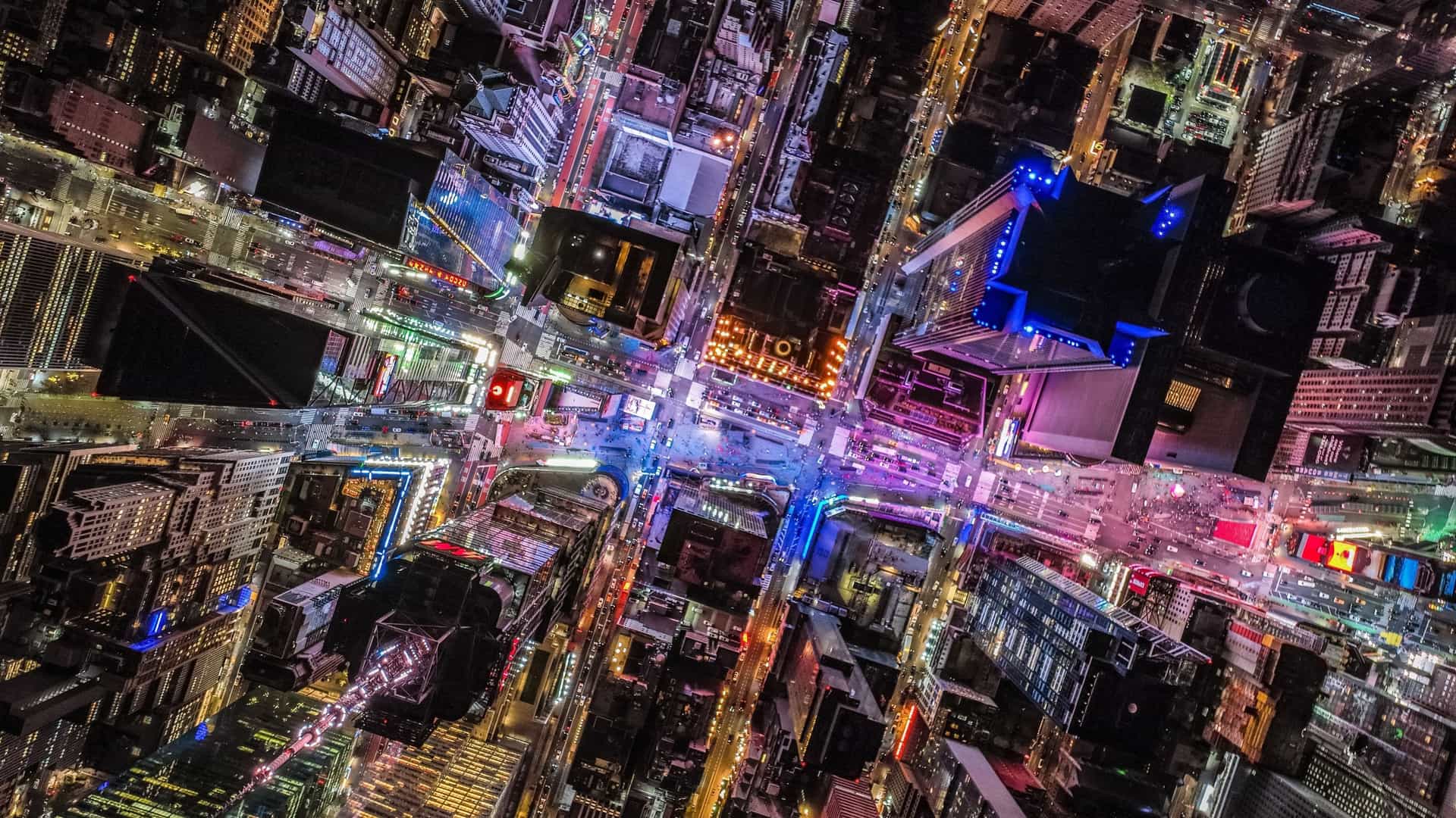 An overhead aerial shot of Times Square in Manhattan, New York City, at night, showing the urban landscape where buildings and streets are saturated with the colorful, glowing lights of massive digital advertisements and video displays.