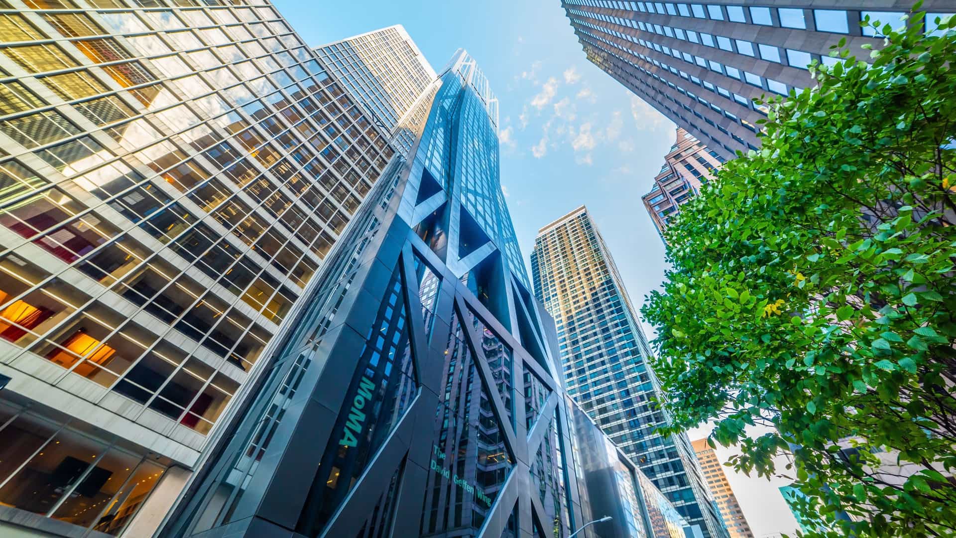 An exterior view of the Museum of Modern Art (MoMA) in New York City. The modern building has a facade of glass and other materials, with the museum's acronym, "MoMA," visible on the building.