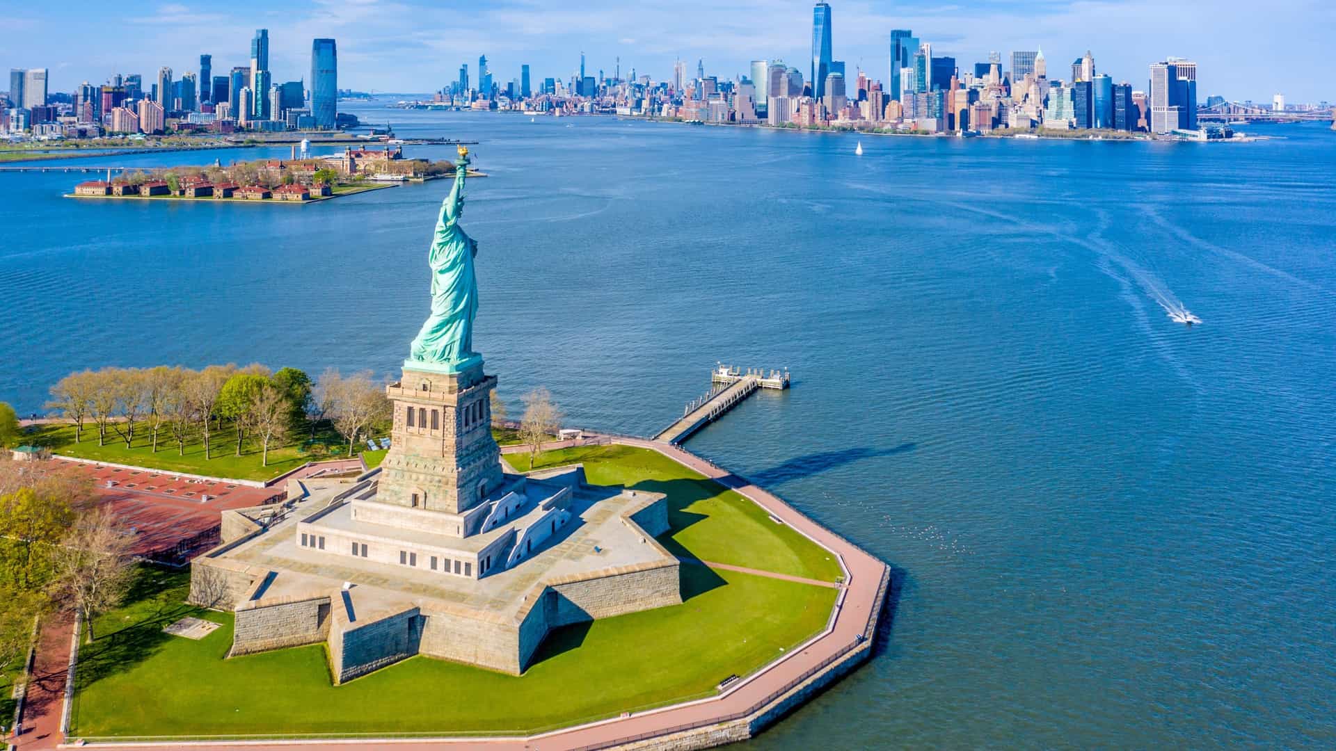An aerial view of the Statue of Liberty and Ellis Island in New York Harbor. The Statue of Liberty is visible on Liberty Island, and Ellis Island is nearby. In the background, the Lower Manhattan skyline is visible.