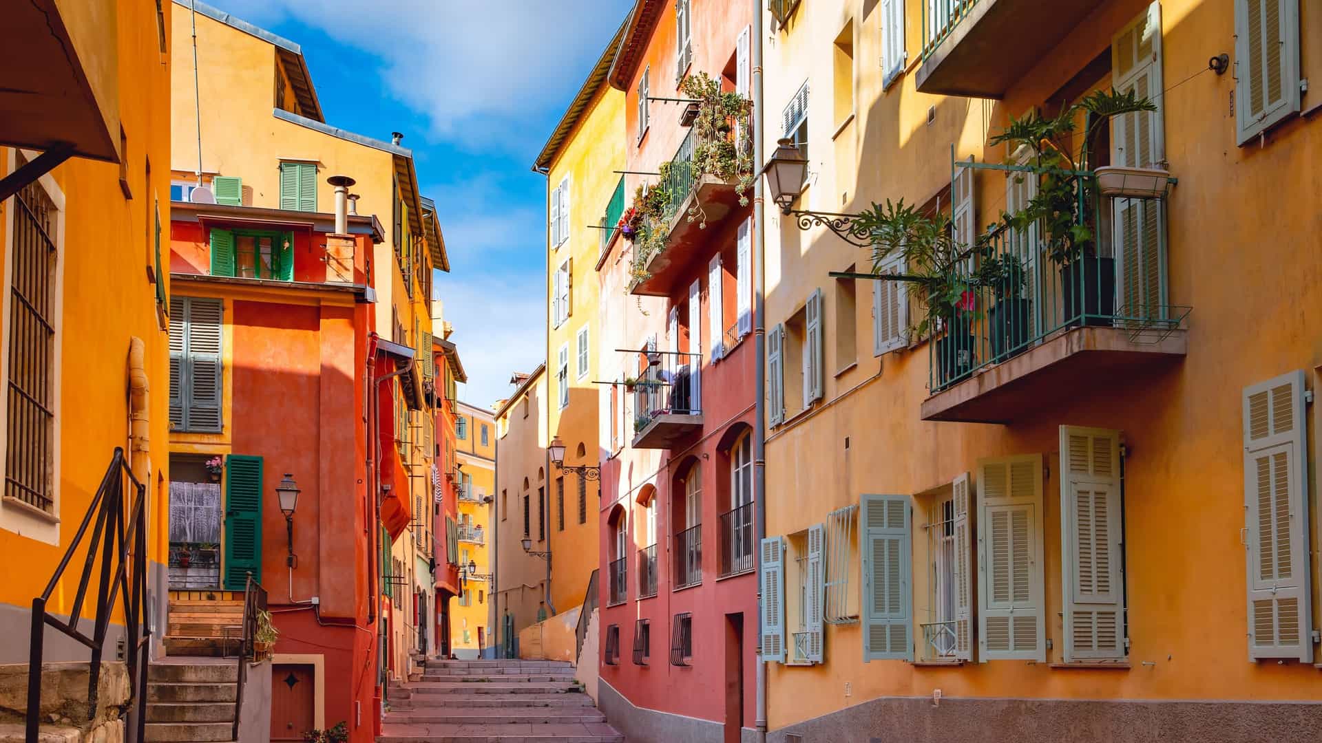 A bright, narrow, sunlit street scene in the Old Town (Vieux Nice) in Nice, France, showing a stepped, cobblestone alleyway lined with historic residential buildings. The facades are painted in vibrant Mediterranean colors, including orange, yellow, and deep terracotta, with windows protected by closed or partially open pale green/blue shutters and small wrought-iron balconies holding potted plants.