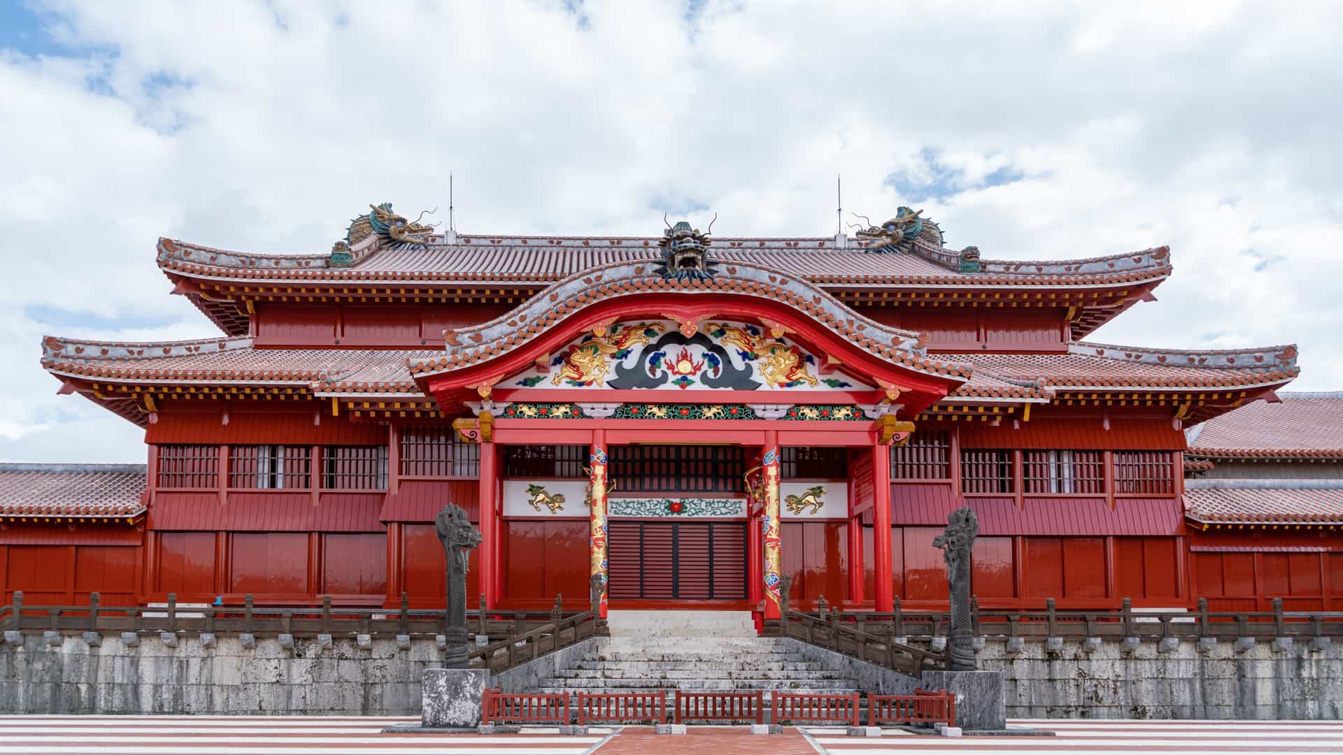 A photograph of the main ceremonial building, the Seiden (main hall), of Shuri Castle in Okinawa, Japan. The impressive, red-tiled structure features a traditional Japanese-Ryukyuan architectural style with wide steps leading up to its main entrance, showcasing the vibrant colors and distinctive corner eaves of the palace grounds.