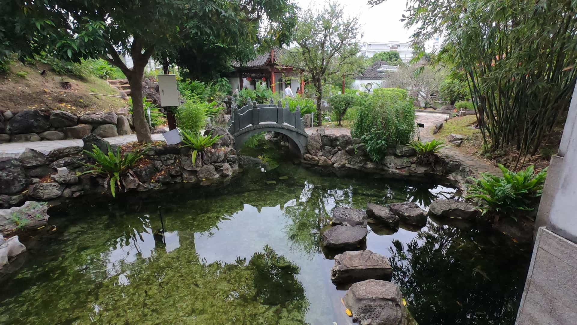 A serene, wide-angle view of the Fukushūen Garden in Naha, Okinawa, Japan. The traditional Chinese-style garden features a large central pond with a reflective surface, surrounded by intricate stone walkways, meticulously shaped trees, and ornate Chinese structures, including a covered bridge and a pavilion with a curved roof.