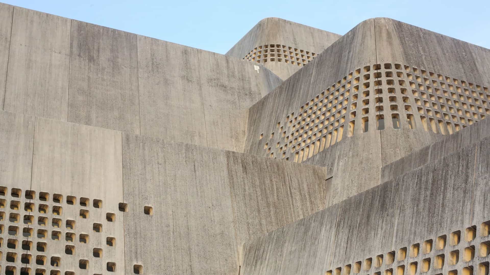 An exterior view of the Okinawa Prefectural Museum & Art Museum in Naha, Japan. The building is a striking example of modern architecture, featuring a massive, fortress-like structure clad in white limestone, designed to resemble traditional Okinawan gusuku (castles). The angular, imposing facade stands against a clear sky, surrounded by landscaped grounds.