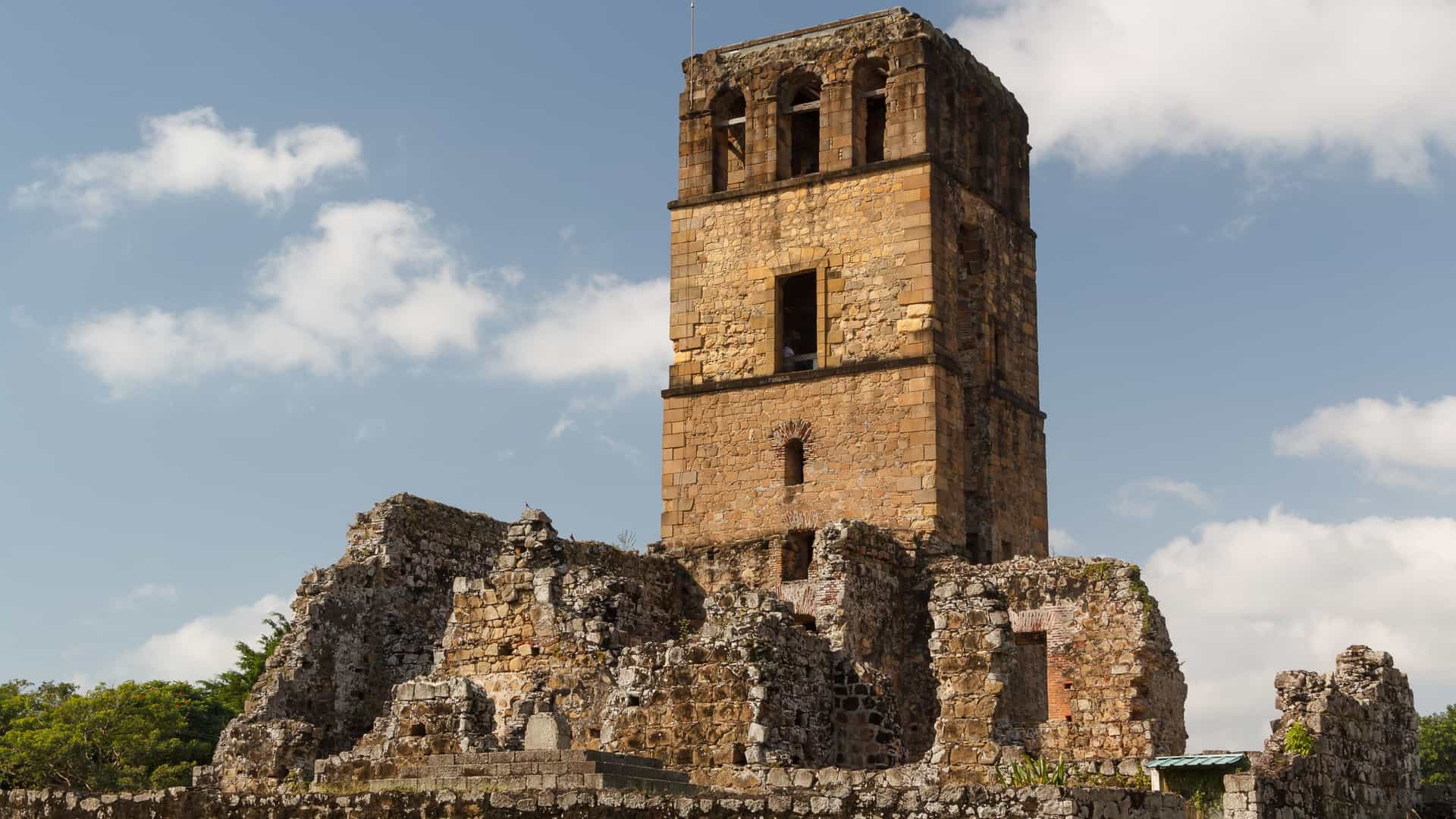  A scenic view of the ruins of Panama Viejo, a UNESCO World Heritage Site in Panama City. The image features the stone ruins of the former colonial city, with the towering, incomplete bell tower of the Cathedral of Our Lady of the Assumption standing prominently in the center. The ruins are surrounded by green grass, and in the distance, the modern, high rise skyline of present day Panama City is visible under a partly cloudy sky, creating a striking contrast between the old and the new.