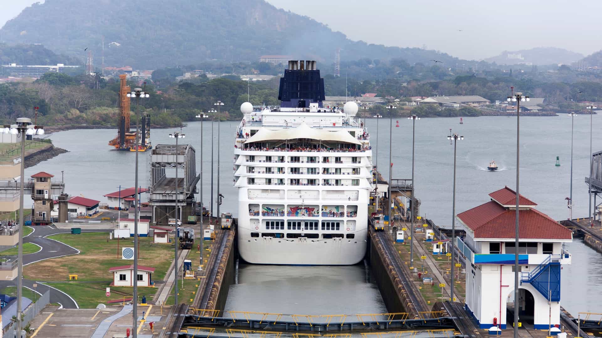  A large cruise ship is shown from a high angle as it navigates the narrow channel of the Panama Canal. The ship is positioned between the massive concrete walls of the Miraflores Locks, with a person visible on the deck near the railing. The water is calm, and the ship appears to be either rising or lowering within the lock, highlighting the impressive engineering of this iconic waterway.