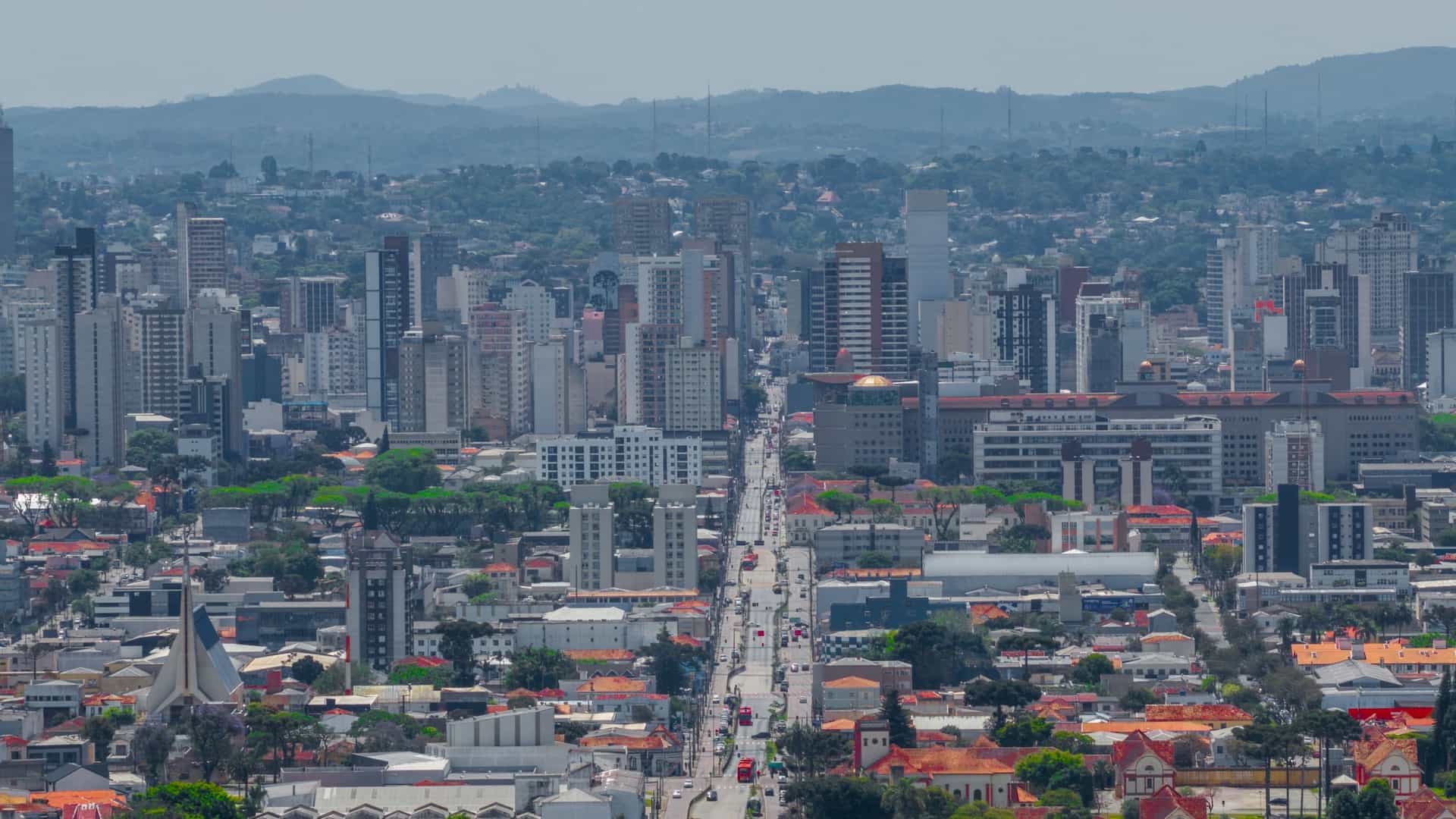 A wide, aerial panoramic view of the dense, modern city center of Curitiba, Brazil, showing a busy avenue cutting through the middle of the frame and lined with high-rise buildings and commercial structures. A distinctive church spire is visible in the foreground, with hazy, undulating mountains visible on the distant horizon.