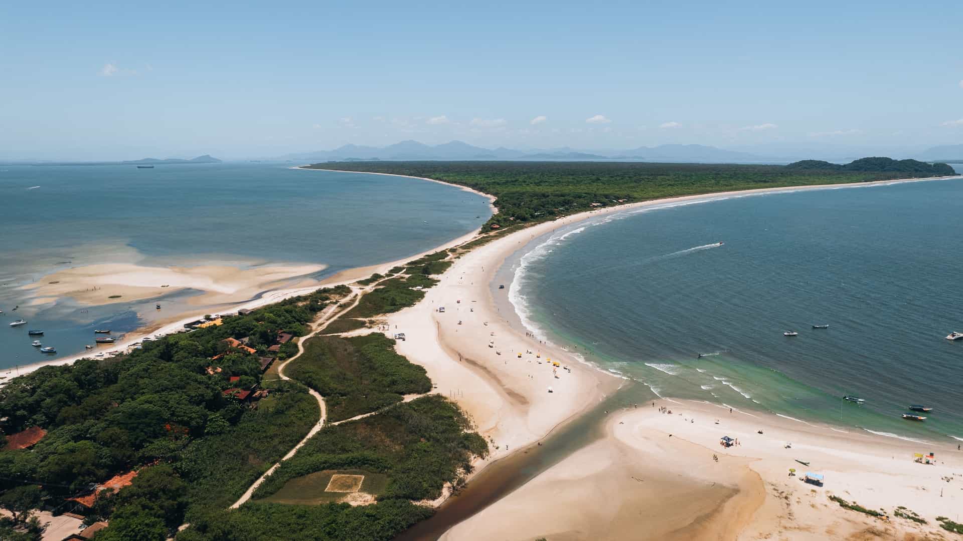 A high-angle aerial view of Ilha do Mel (Honey Island), Paraná, Brazil. The image shows a narrow, sandy isthmus of land connecting two larger forested sections, with the white, slender Farol das Conchas (Conchas Lighthouse) visible atop a small, green hill near the point where the isthmus widens. Turquoise water and white-sand beaches surround the lush Atlantic Rainforest vegetation.
