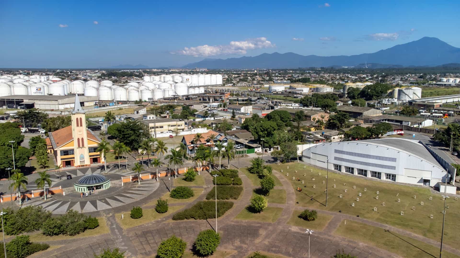 An aerial view of the Catedral Nossa Senhora do Rosário in Paranaguá, Paraná, Brazil. The historic cathedral features a large central body with a white facade and a tall, slender Neo-Gothic bell tower standing against a backdrop of the city's denser urban buildings, with the Paranaguá Bay and docks visible in the distance.
