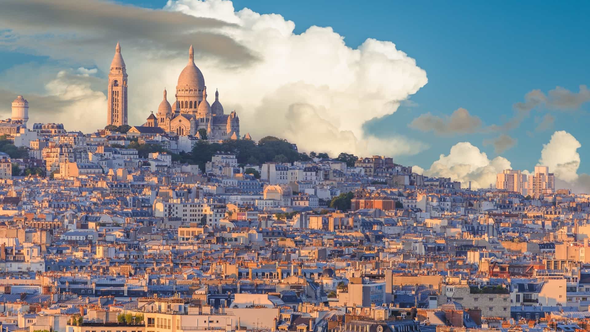  A panoramic view of Paris from the Montmartre hill, with the white domes and bell tower of the Basilica of the Sacré Cœur visible in the foreground. The basilica stands out against the backdrop of the sprawling Parisian cityscape under a bright sky.