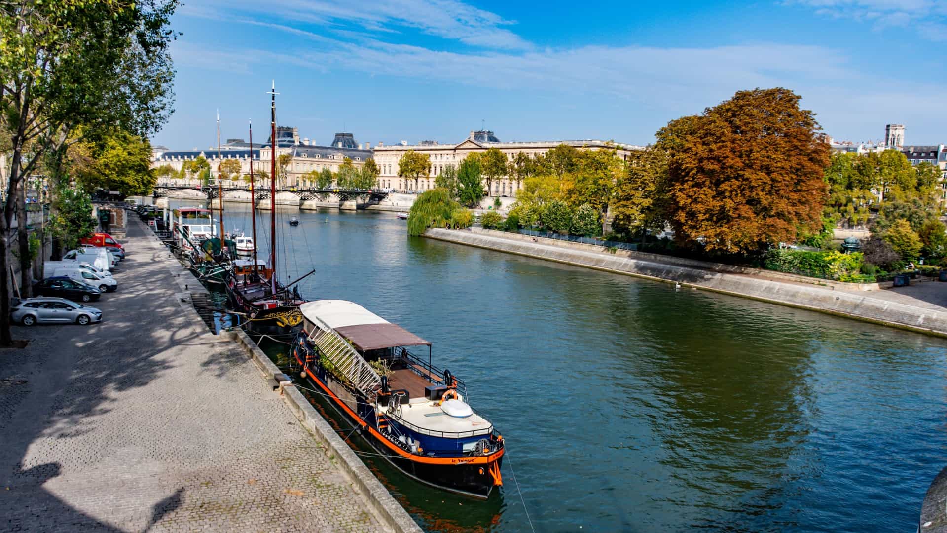  Barges and other boats are moored along the banks of the Seine River in Paris. The river is filled with various vessels, including tourist boats and residential barges, against a backdrop of a bridge and city buildings. The scene depicts a typical day on the river.
