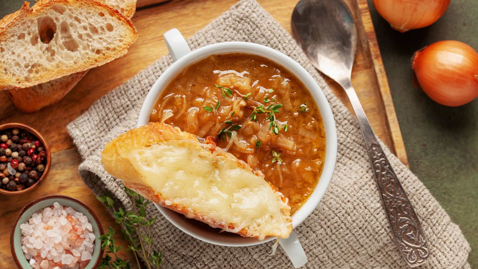 A close-up shot of classic French Onion Soup (Soupe à l'oignon gratinée) served in a traditional, handled ceramic crock. A thick, golden-brown crust of melted and baked cheese (likely Gruyère) completely covers the top of the soup, sealing in the savory broth and caramelized onions. The edges of the cheese are bubbling and slightly caramelized from being recently broiled.