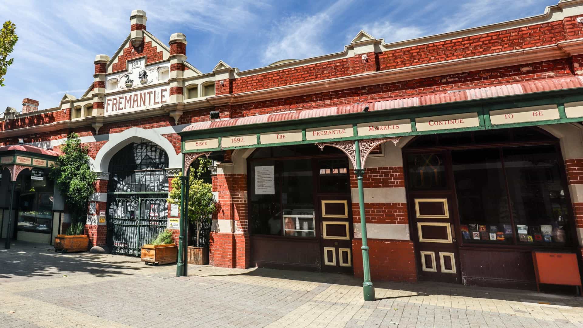 A close-up view of the historic, Federation-style facade of the Fremantle Markets building. The long structure is made of brick and features large, arched entrances and many windows, all sheltered under a prominent, dark-colored corrugated roof. The market name is prominently displayed above the central archway, set against a blue sky with some white clouds.