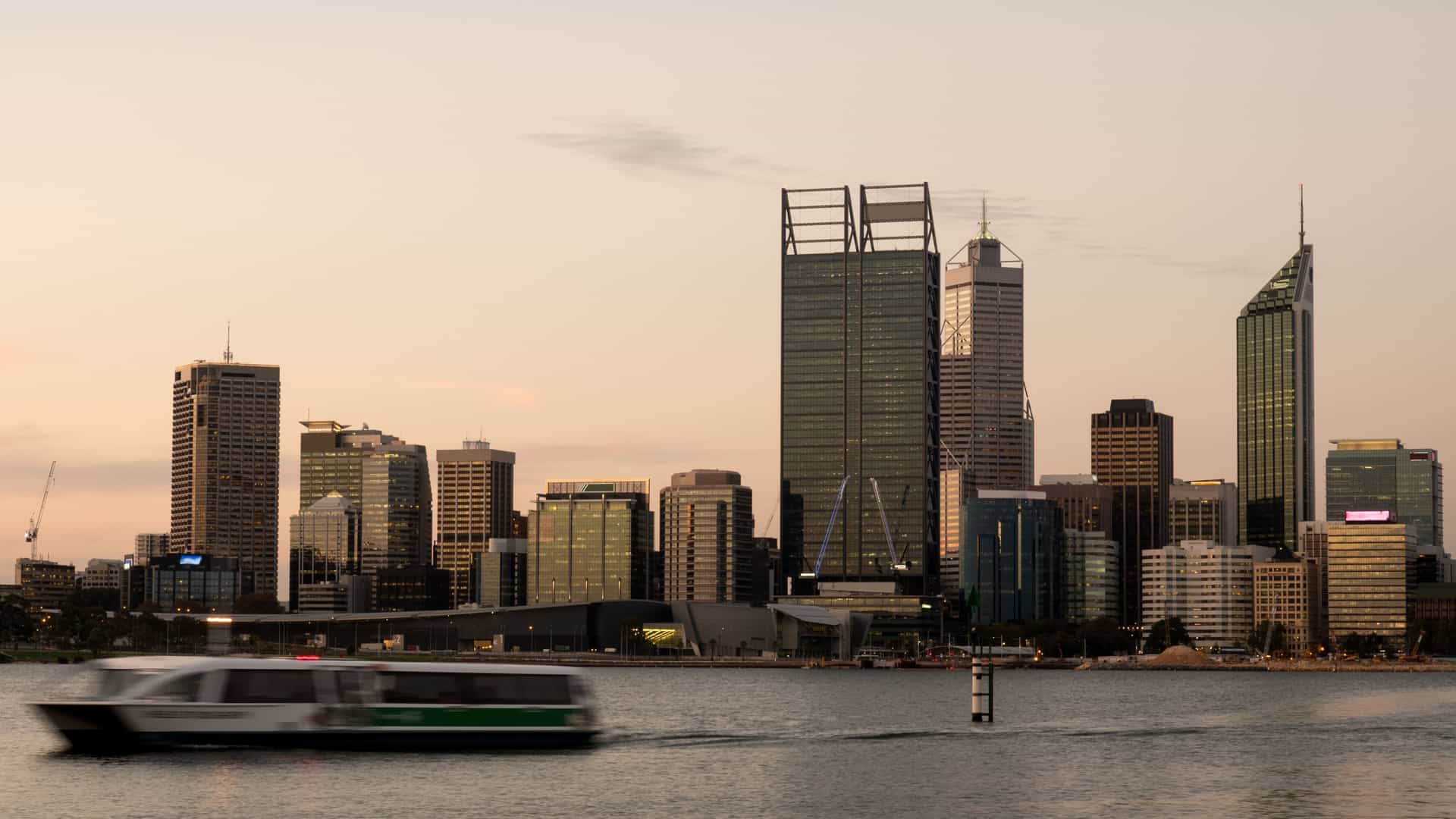 A blue and white passenger ferry travels across the calm waters of the Swan River in Perth, Western Australia. The boat creates a wake behind it. In the background, the dense, modern skyline of the city, including numerous high-rise buildings, rises beneath a mostly clear sky.