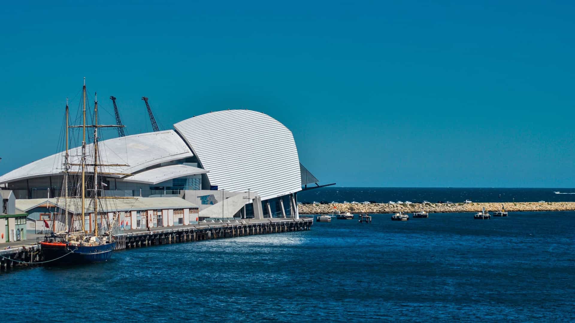 A bright, modern view of the angular, white facade of the Western Australian Maritime Museum building in Fremantle, situated right next to the water of the harbor. The long, multi-level building features a curved roofline that dramatically sweeps downward toward the water. In the foreground, there are grassy areas and paved pathways leading up to the museum entrance.