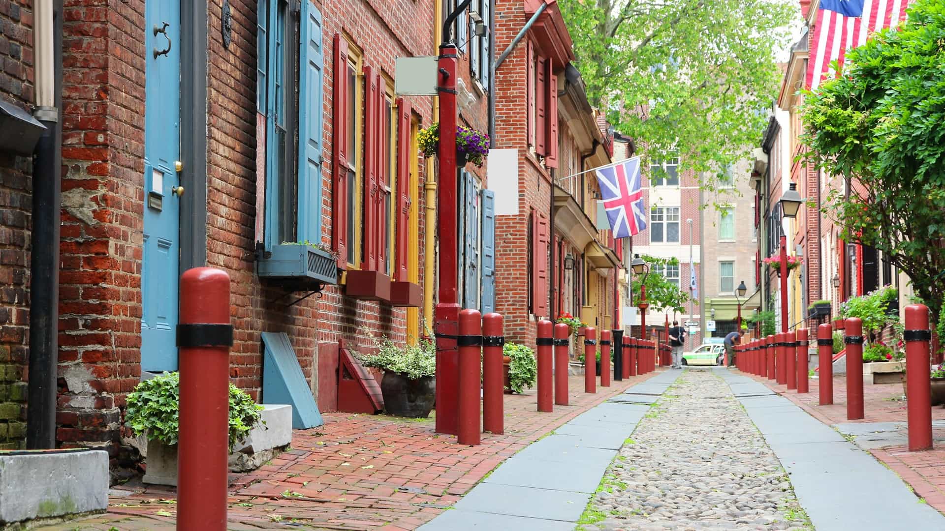  A view down Elfreth's Alley, a narrow, historic cobblestone street in Philadelphia. The alley is lined with colorful, colonial era brick row houses with shutters, many of which have flower boxes. The street appears to be empty, giving a quiet, historical atmosphere.