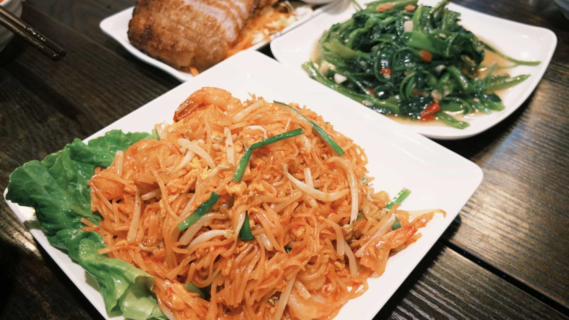 A close-up, appetizing flat-lay photograph showcasing a diverse spread of classic Thai cuisine. Featured dishes include plates of Pad Thai, bright green and yellow curries, stir-fried vegetables, and bowls of soup, all garnished with fresh herbs and lime wedges. The collection of dishes highlights the colorful and complex flavors of Thai cooking.