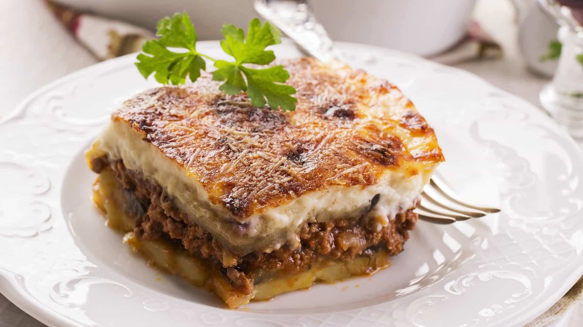 A close-up, top-down photograph of a square serving of baked Moussaka on a white plate. The dish consists of layers of sliced eggplant, ground lamb (or beef), and potatoes, topped with a thick, golden-brown baked layer of creamy béchamel sauce. The edges show the separation of the layered ingredients.
