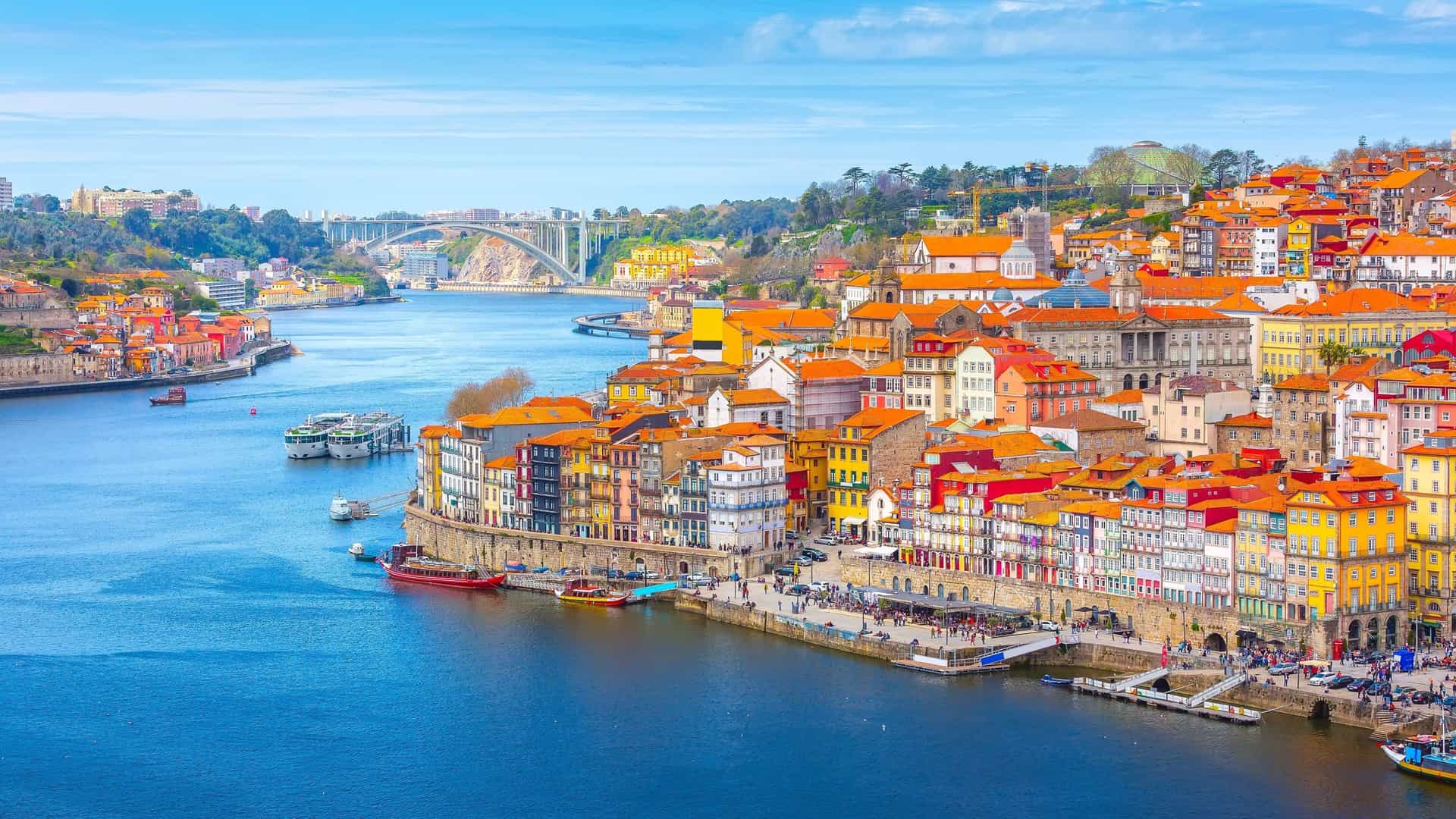 Aerial view of Porto's historic Ribeira district, featuring the colorful, tightly packed houses along the promenade of the Douro River, with traditional Rabelo boats docked in the foreground under a clear blue sky.