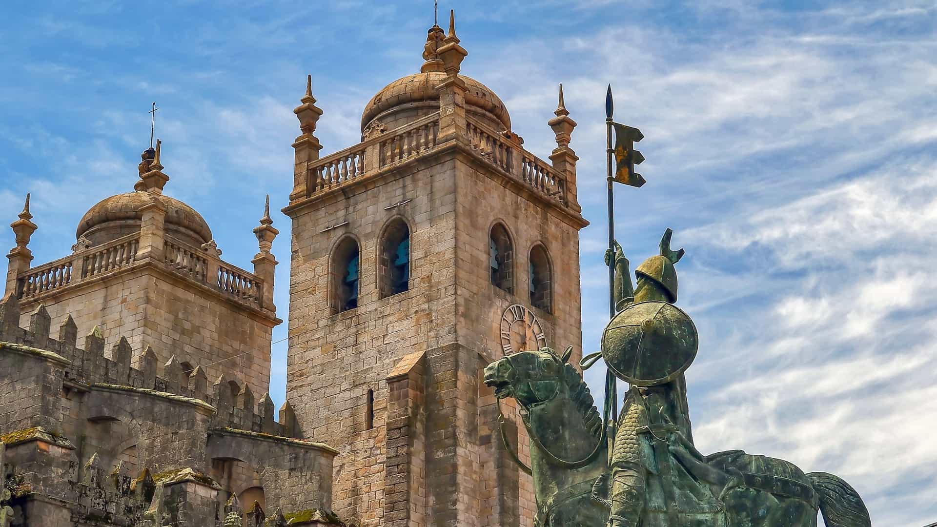 A view of the fortress like Porto Cathedral (Sé do Porto) in Portugal, a Romanesque landmark with twin towers, standing behind the equestrian statue of Vimara Peres in the Terreiro da Sé plaza on a bright day.