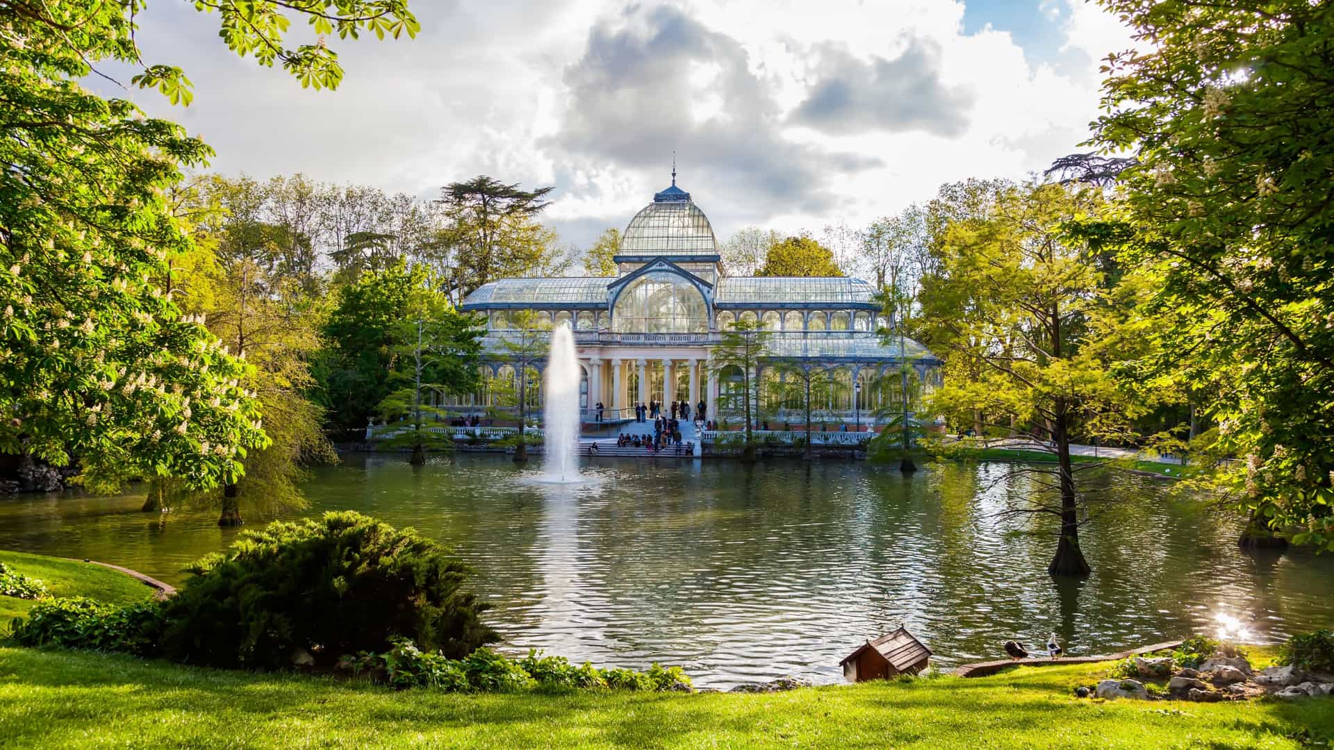The Crystal Palace (Palacio de Cristal), a stunning glass and iron conservatory in Retiro Park, Madrid, Spain. The structure stands by a small pond with lush green surroundings on a bright, sunny day.