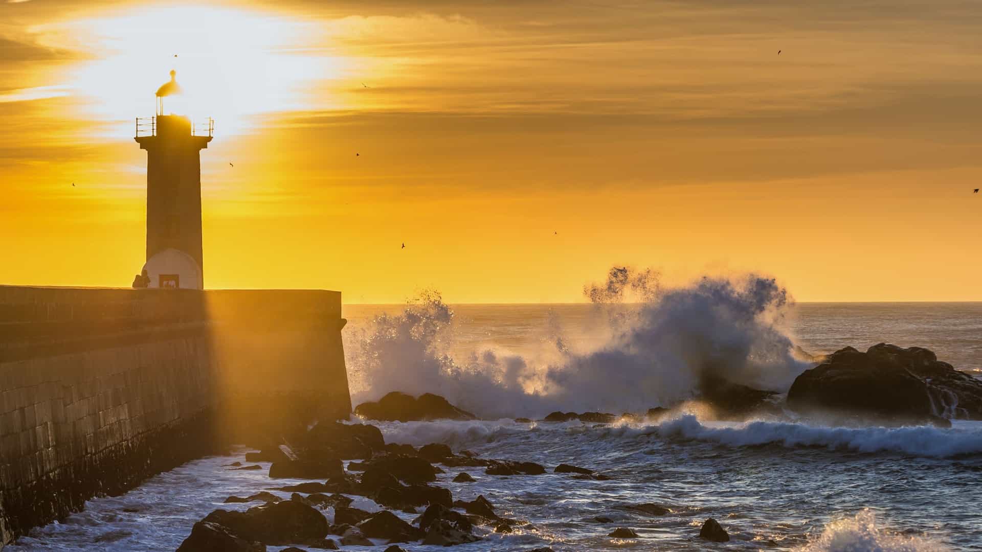 Felgueiras Lighthouse (Farol de Felgueiras) on the Atlantic coast of Porto, Portugal, silhouetted against a dramatic sunset sky. The stone breakwater is battered by large ocean waves.