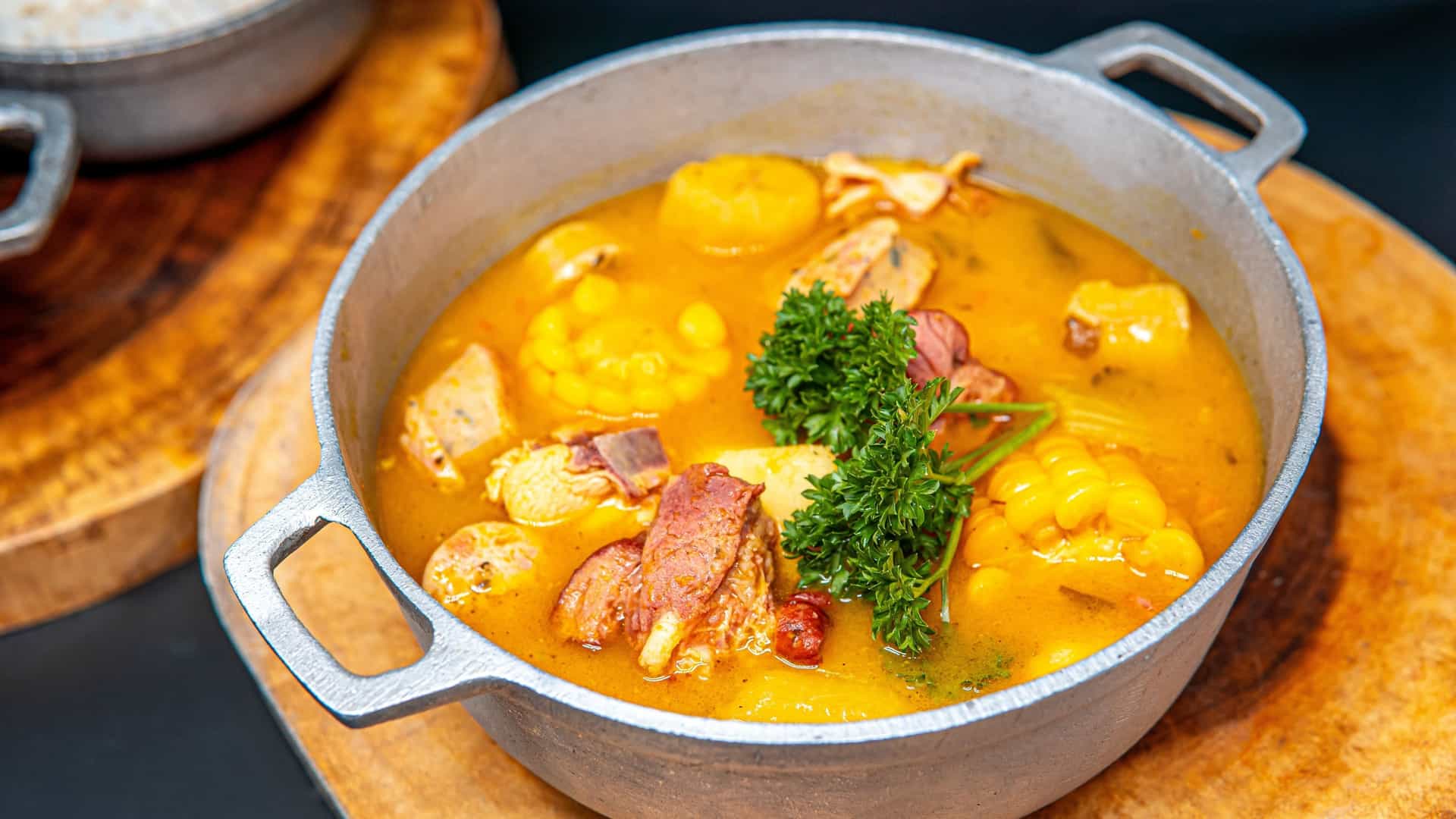 A close-up, appetizing photograph of a large bowl of Sancocho, a flavorful, hearty Dominican stew. The rich, dark broth is filled with various chunks of meat, corn cobs, and root vegetables like plantain and yuca. The stew is garnished with herbs and served with a side of white rice and slices of avocado, illustrating a traditional Dominican meal.