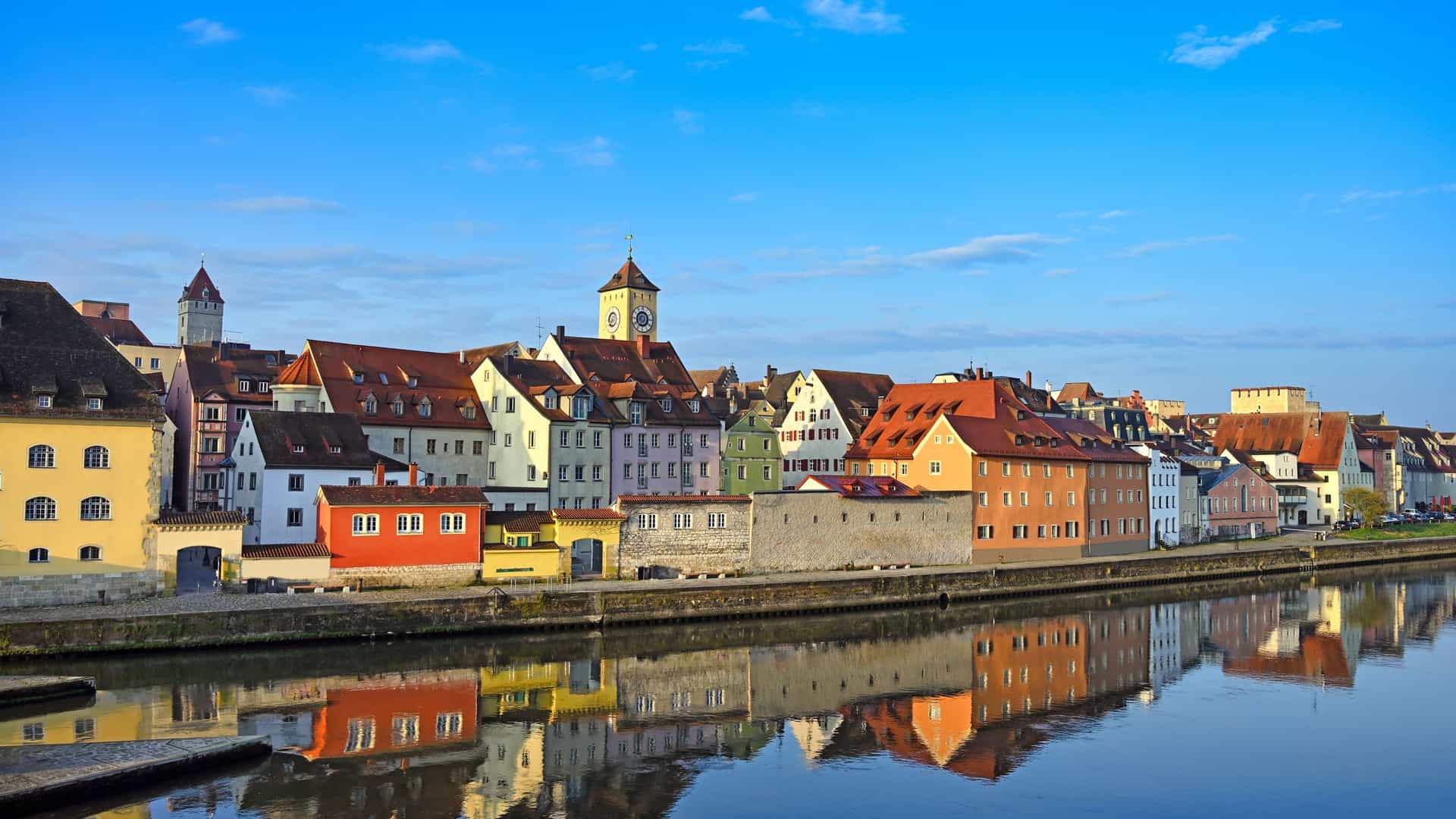  A picturesque panoramic view of Regensburg's UNESCO Old Town, featuring colorful buildings and the historic Stone Bridge spanning the tranquil Danube River in Bavaria.