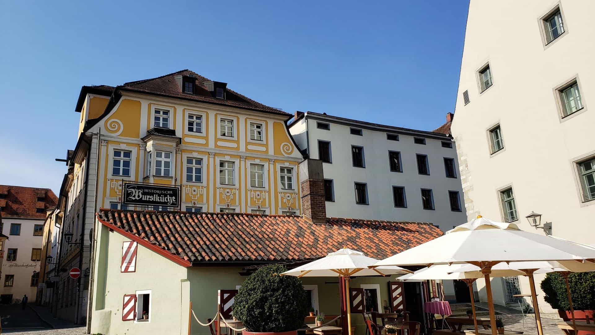 The outdoor seating area of the historic Wurstkuchl (Oldest Sausage Kitchen) in Regensburg, Germany. The scene features tables with umbrellas on a cobblestone street, next to a small green building and a larger yellow historic structure.