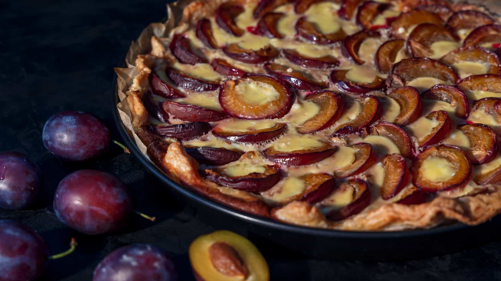 A close-up photograph of a homemade Quetschentaart, a traditional plum tart from the Luxembourg Moselle Valley. The tart features deep purple plums (Quetsches) baked into a pastry base with a light custard filling, surrounded by whole, ripe plums on a dark surface.