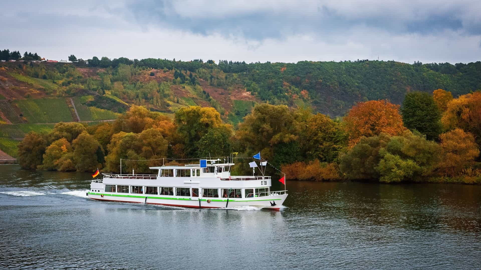 A river cruise boat carrying passengers sails along the wide, calm Moselle River in Germany. The banks of the river are lined with lush, colorful autumn foliage and forested hillsides, with a mix of vineyards visible on the slopes in the distance.
