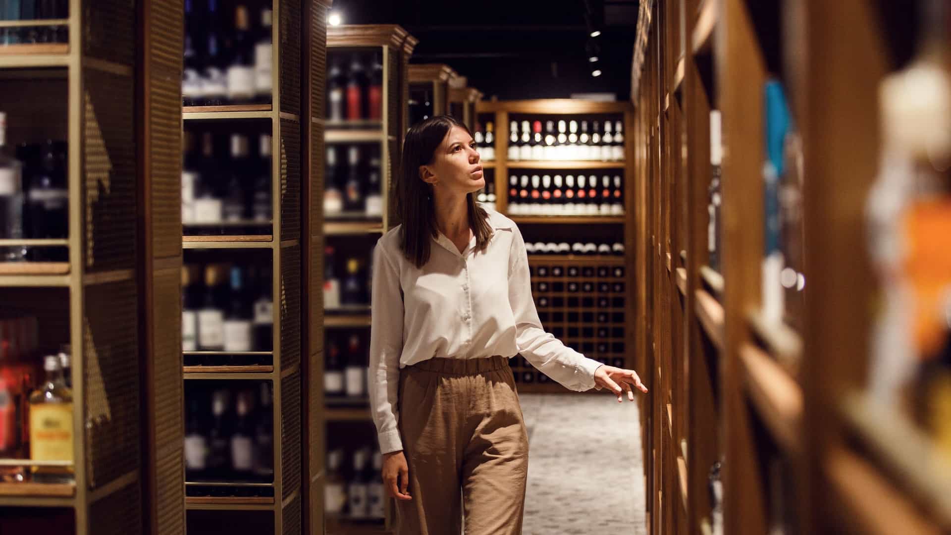 A young woman in a white shirt and brown trousers stands in the dimly lit aisle of a sophisticated, wood-paneled wine shop or cellar, looking up at bottles stacked on tall shelves. This image perfectly captures the experience of a visitor who wants to (shop for wines in remich luxemberg for a souvenir).