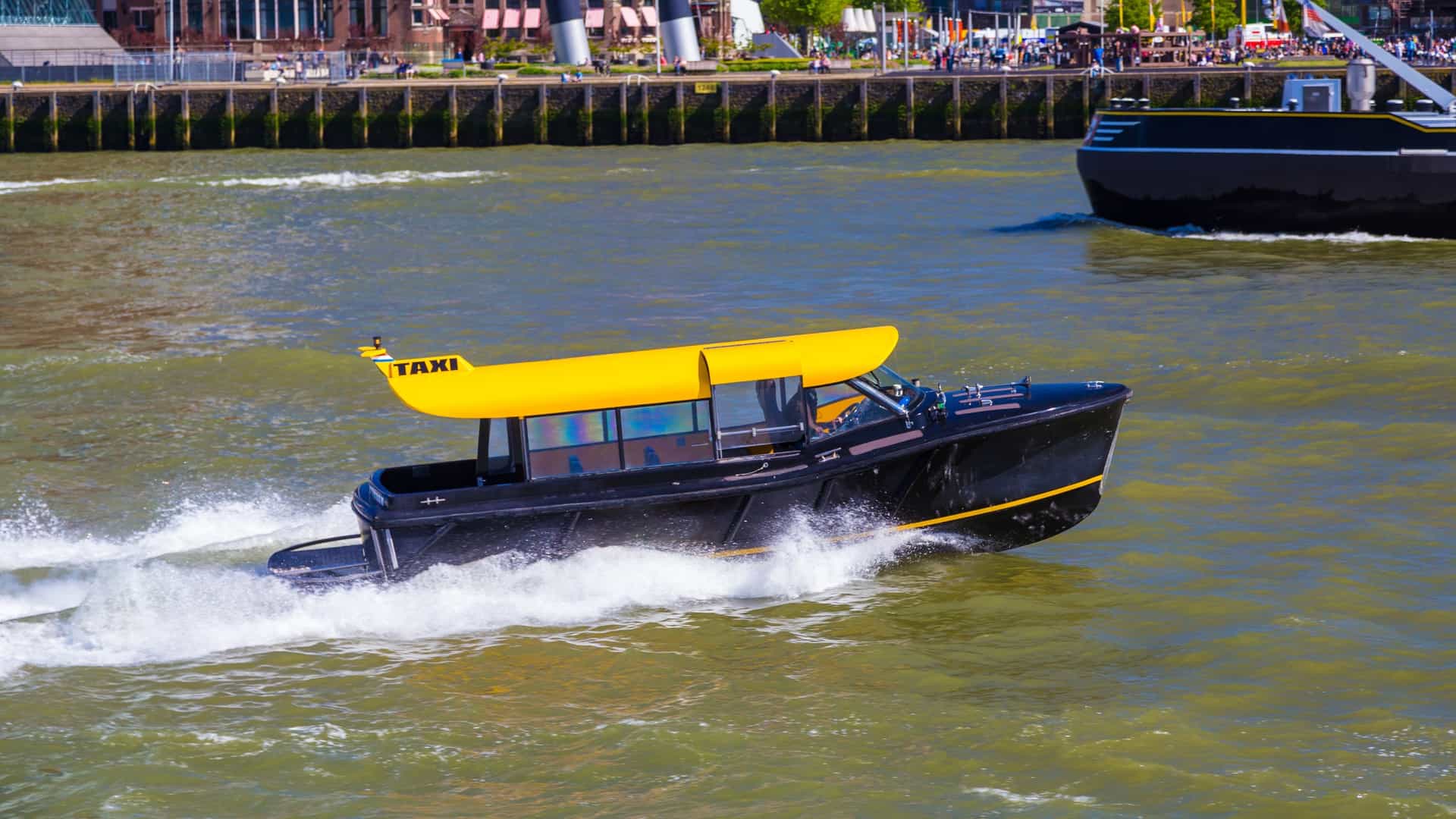 A black and yellow high-speed Water Taxi cuts across the Nieuwe Maas River in Rotterdam, Netherlands, leaving a white wake behind it. In the background, the modern skyline of Rotterdam is visible under a slightly overcast sky.
