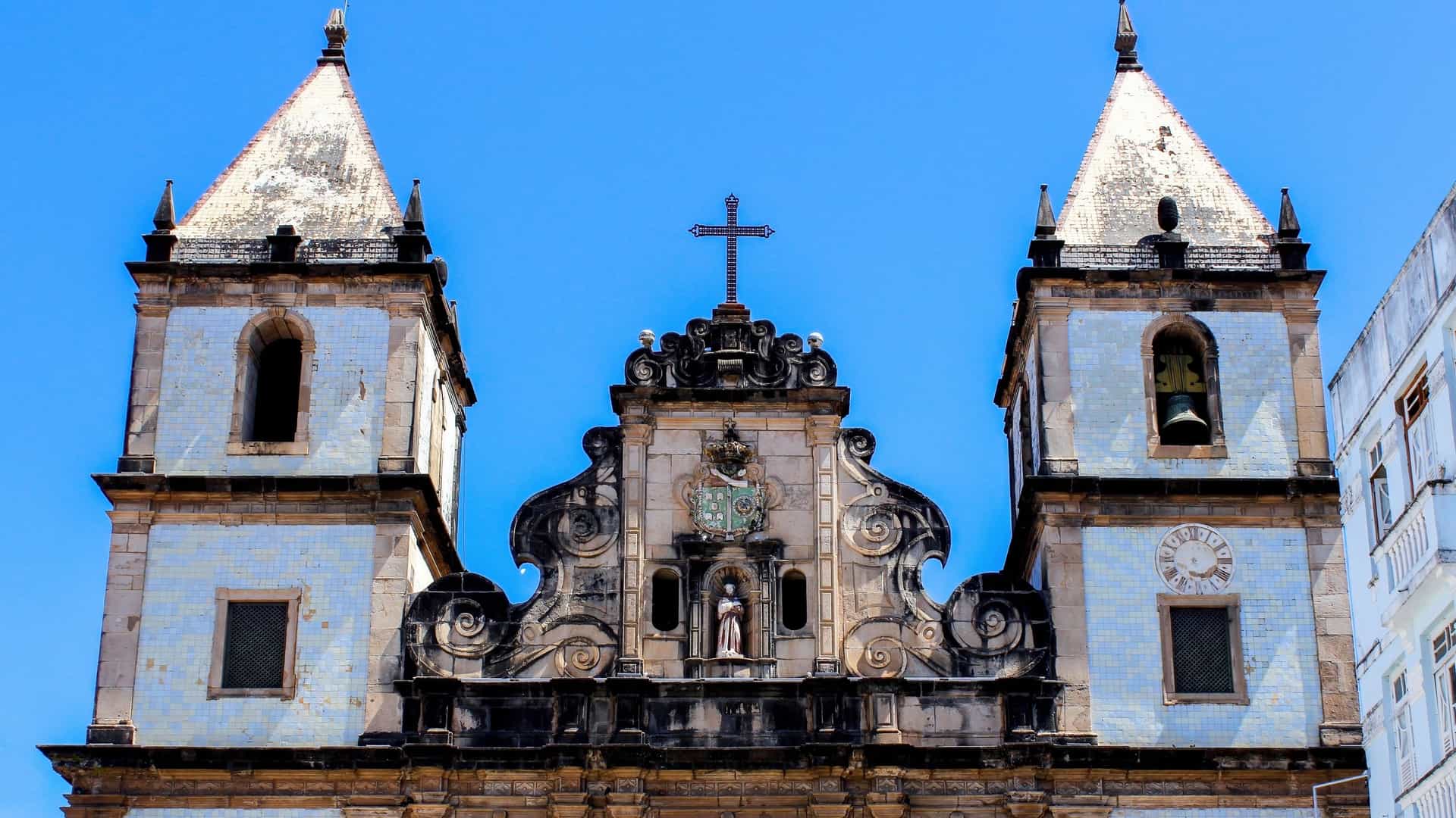 An exterior view of the Church and Convent of São Francisco in Salvador, Bahia, Brazil. The Baroque stone façade features three large entrances and two flanking bell towers, standing on the edge of the historic Pelourinho district, often seen with a large stone cross in the foreground square.