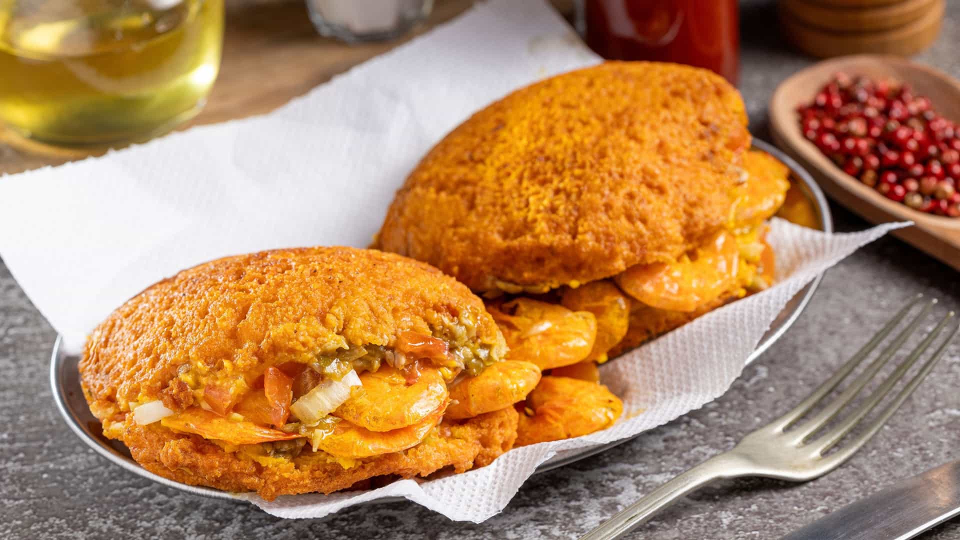 A close-up photograph captures Acarajé, an iconic and traditional Afro-Brazilian street food from Bahia, presented with rich, vibrant fillings. The deep-fried golden fritter, made from black-eyed peas and fried in dendê oil, is split open and generously stuffed with creamy, yellow vatapá and the green, okra-based caruru paste, topped with savory dried shrimp.