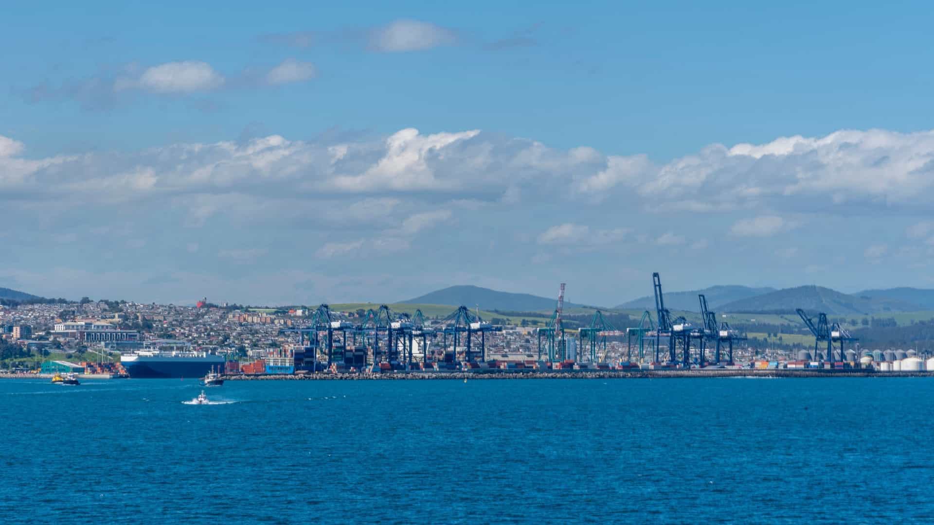 A sweeping panoramic photograph of San Antonio, Chile, on a sunny day captures the busy industrial coastline and harbor. The vast Pacific Ocean dominates the foreground, where multiple cargo container ships are anchored or moving, underscoring the city's importance as Chile's primary freight port. The city's dense urban area, featuring various buildings and infrastructure, climbs the hills behind the port's industrial piers and towering gantry cranes, all under a clear blue sky.