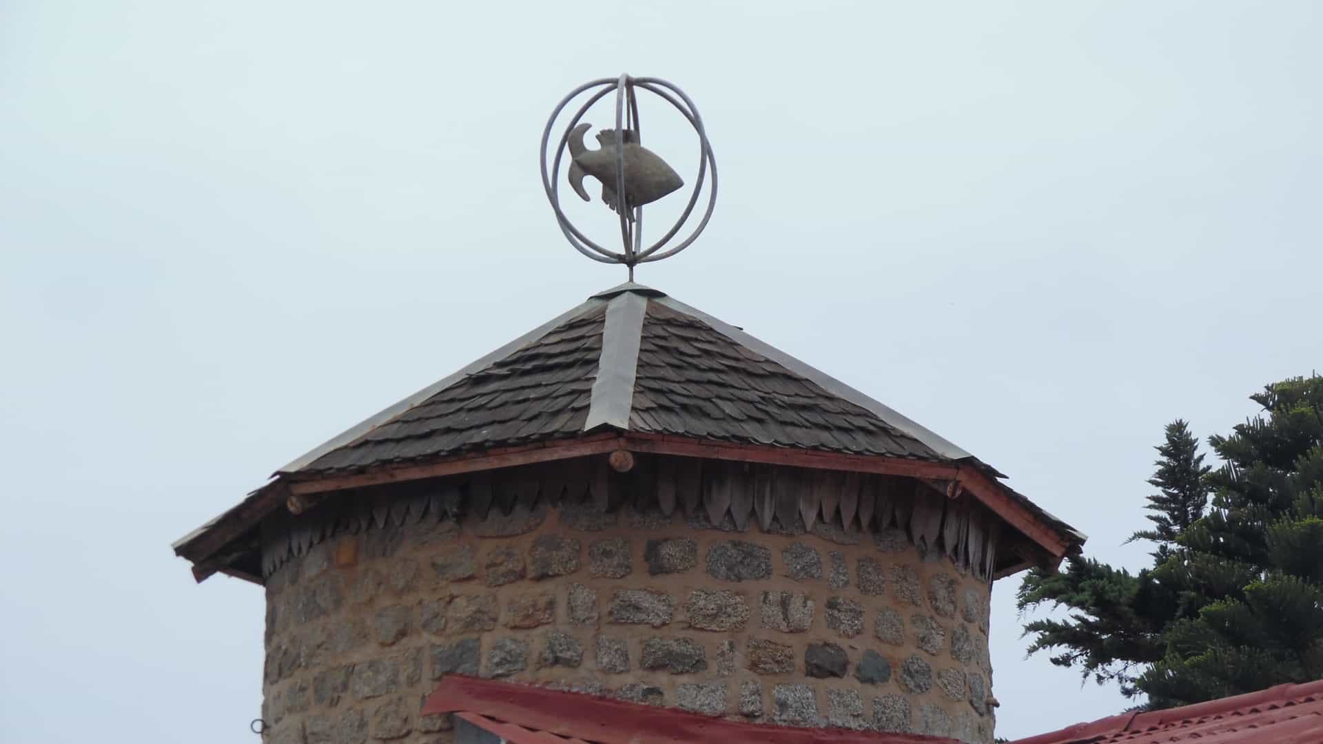 A wide view of the Casa de Pablo Neruda at Isla Negra, Chile, showcasing the poet's unique, ship-like coastal home constructed with stone, wood, and galvanized metal roofing, situated high above the Pacific Ocean's rugged, dark rock shoreline.
