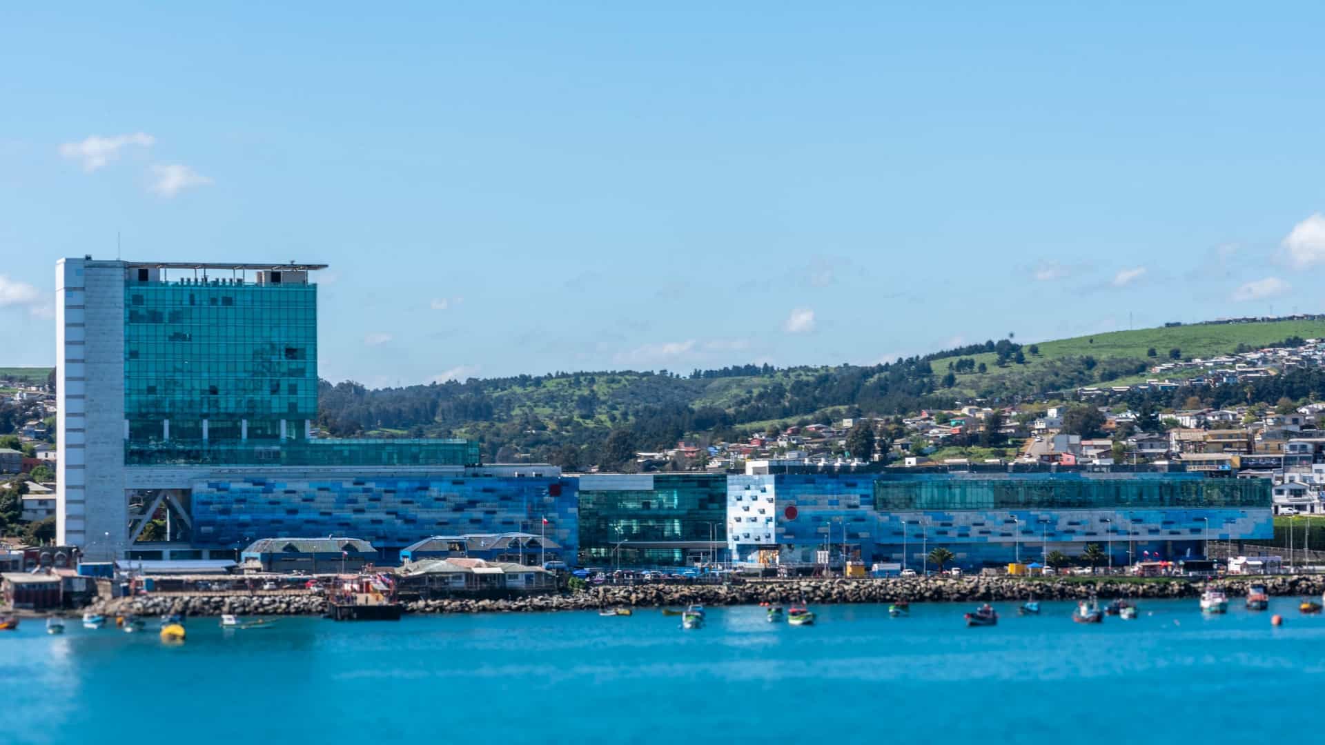 A horizontal, daylight image showing the modern waterfront of the town of San Antonio, Chile. The foreground features the bright turquoise water of a small harbor with several fishing boats anchored behind a rock breakwater. Dominating the center is a large, contemporary building complex with a striking, blue-tinted glass facade and a tall, multi-story tower. The cityscape extends toward the background, where the city's residential areas climb the adjacent green hills under a bright blue sky with sparse white clouds.